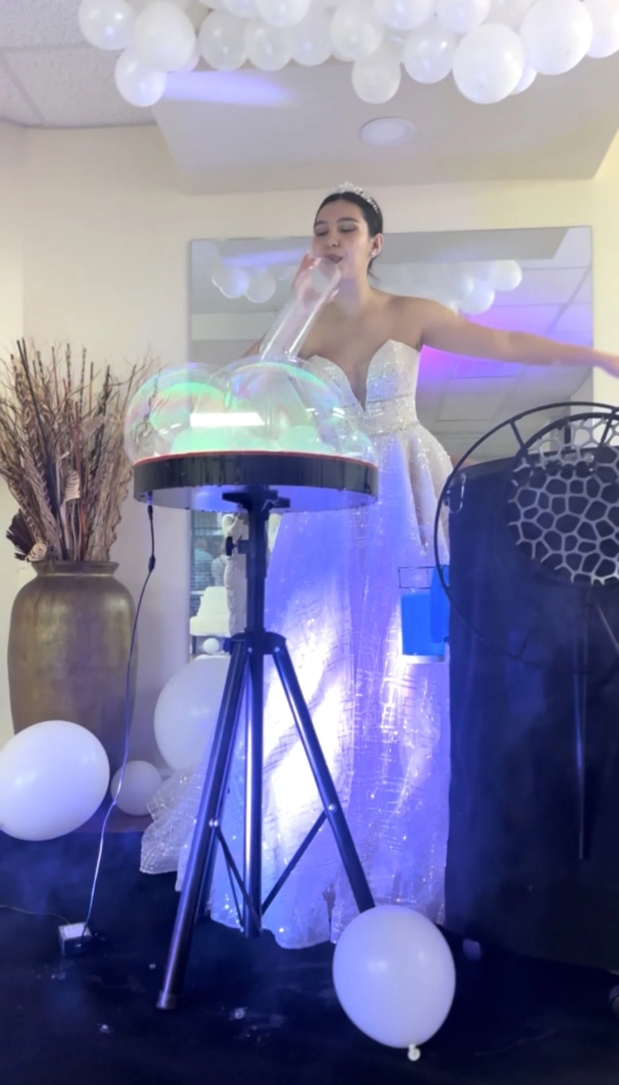 A bride in a white wedding gown and tiara is performing a scientific experiment or demonstration involving a large glass test tube and a container of liquid, with decorative balloons and a vase with dried flowers in the background.
