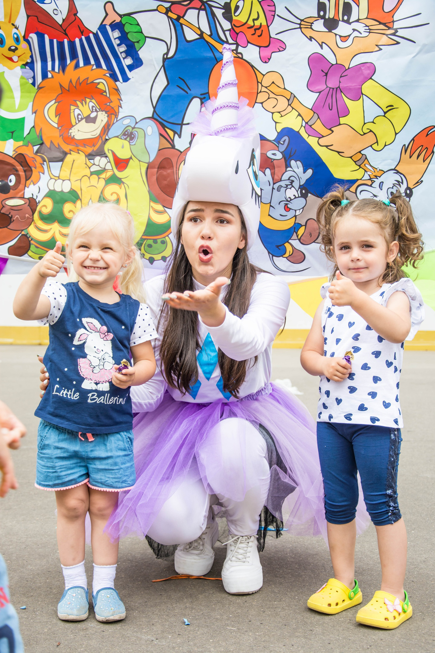 A woman dressed as a unicorn blowing bubbles with two young girls, one giving a thumbs up and the other with a thumbs up, in front of a colorful carnival or festival backdrop featuring cartoon characters.