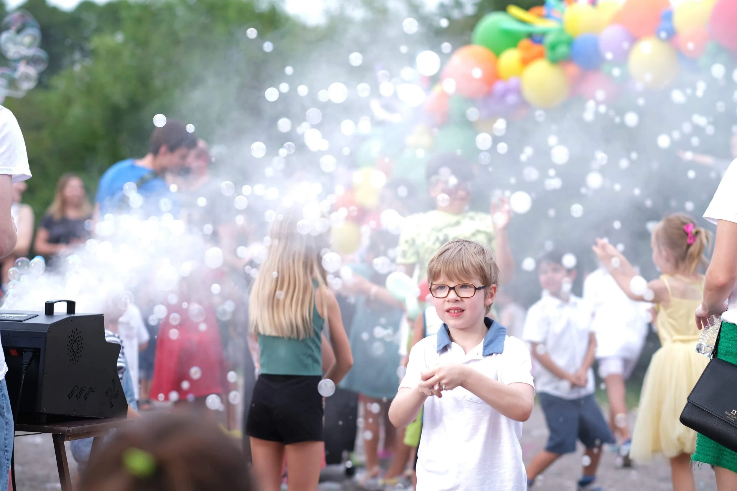 Children at a celebration enjoying a bubble machine with bubbles floating in the air. Some children are looking at the camera while others are playing with bubbles. In the background, there are colorful balloons and trees.