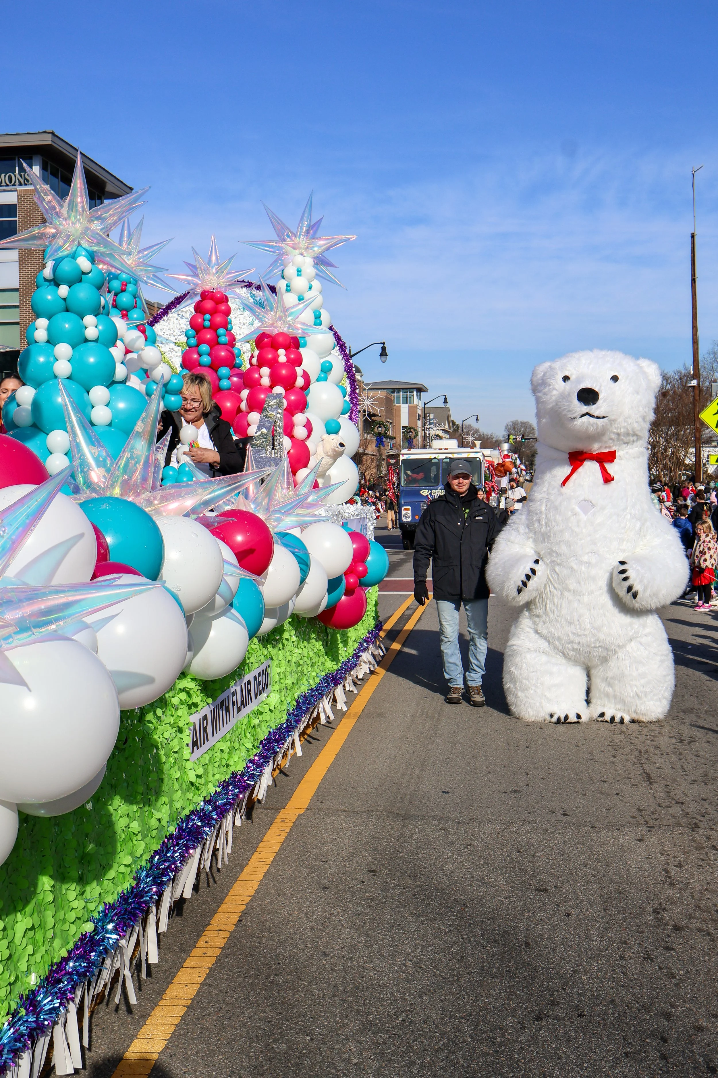 Festive parade float decorated with blue, pink, and white balloons, star-shaped ornaments, and greenery, with a person in a polar bear costume and a person in a black jacket walking beside it on a sunny street filled with onlookers.