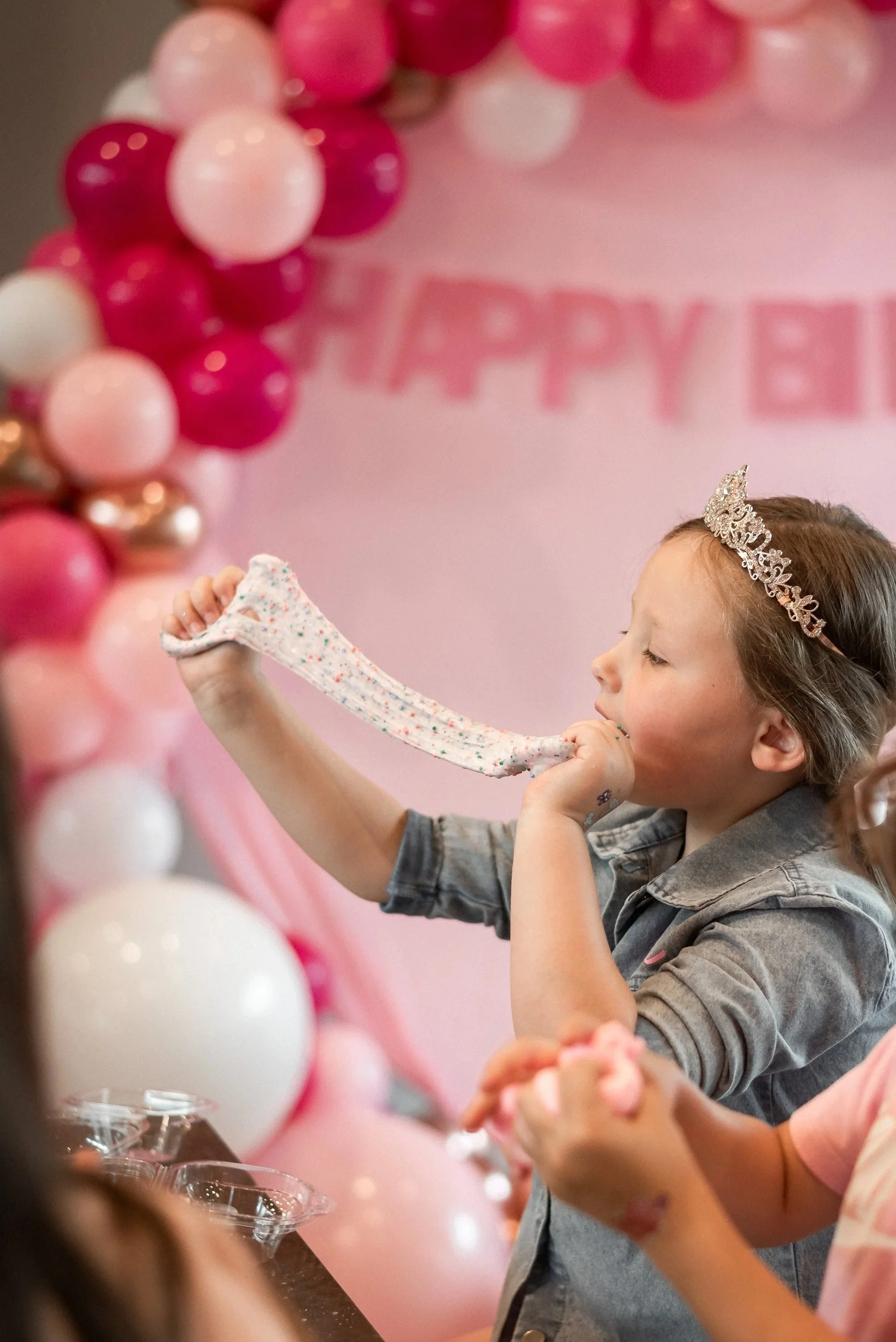 A young girl with a tiara on her head, blowing up a long, colorful balloon in front of a pink and white balloon decorated backdrop with a partially visible sign that reads 'HAPPY B'.