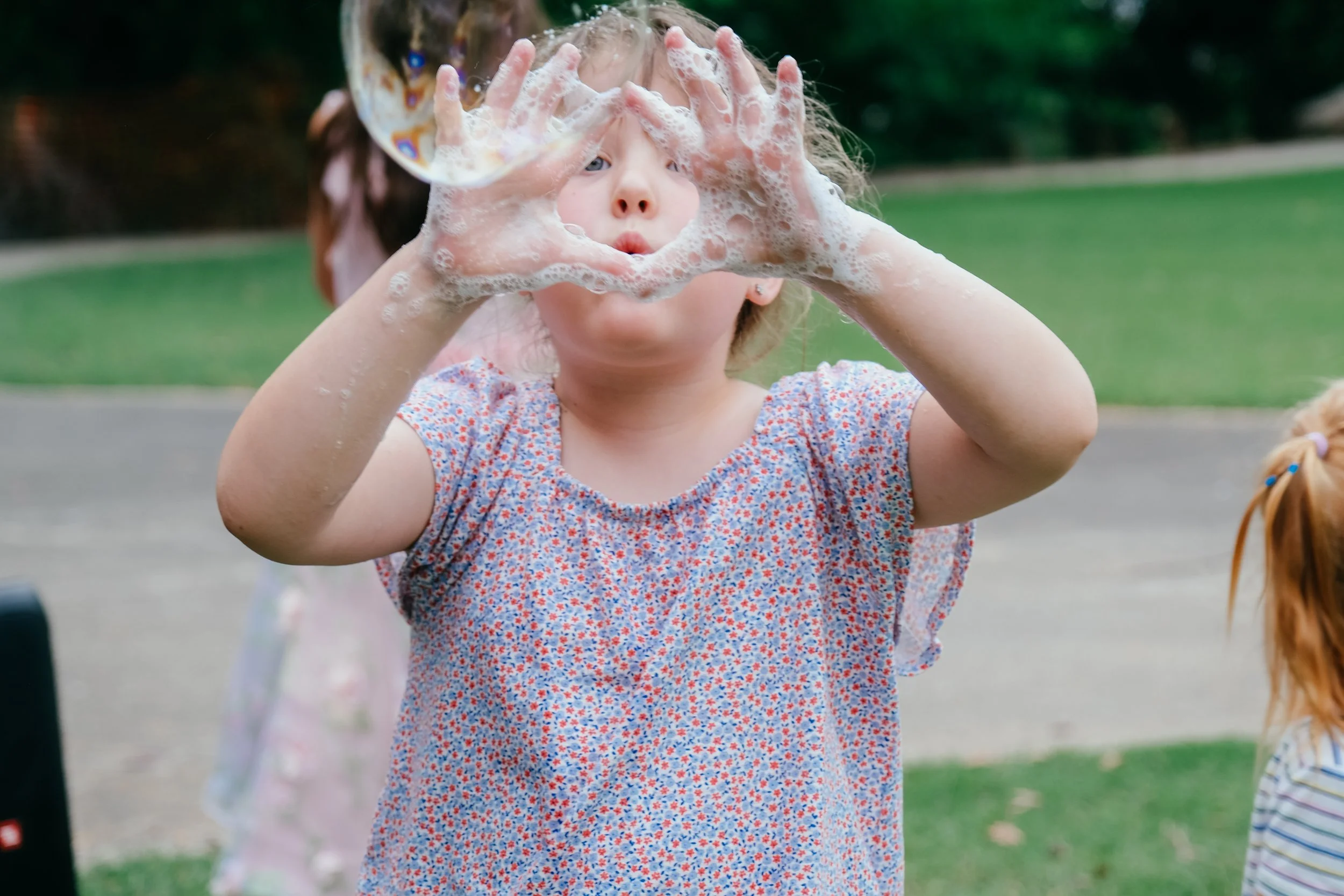 A young girl with short blonde hair and a floral shirt is playing outdoors with soap bubbles, holding foam-covered hands in front of her face.