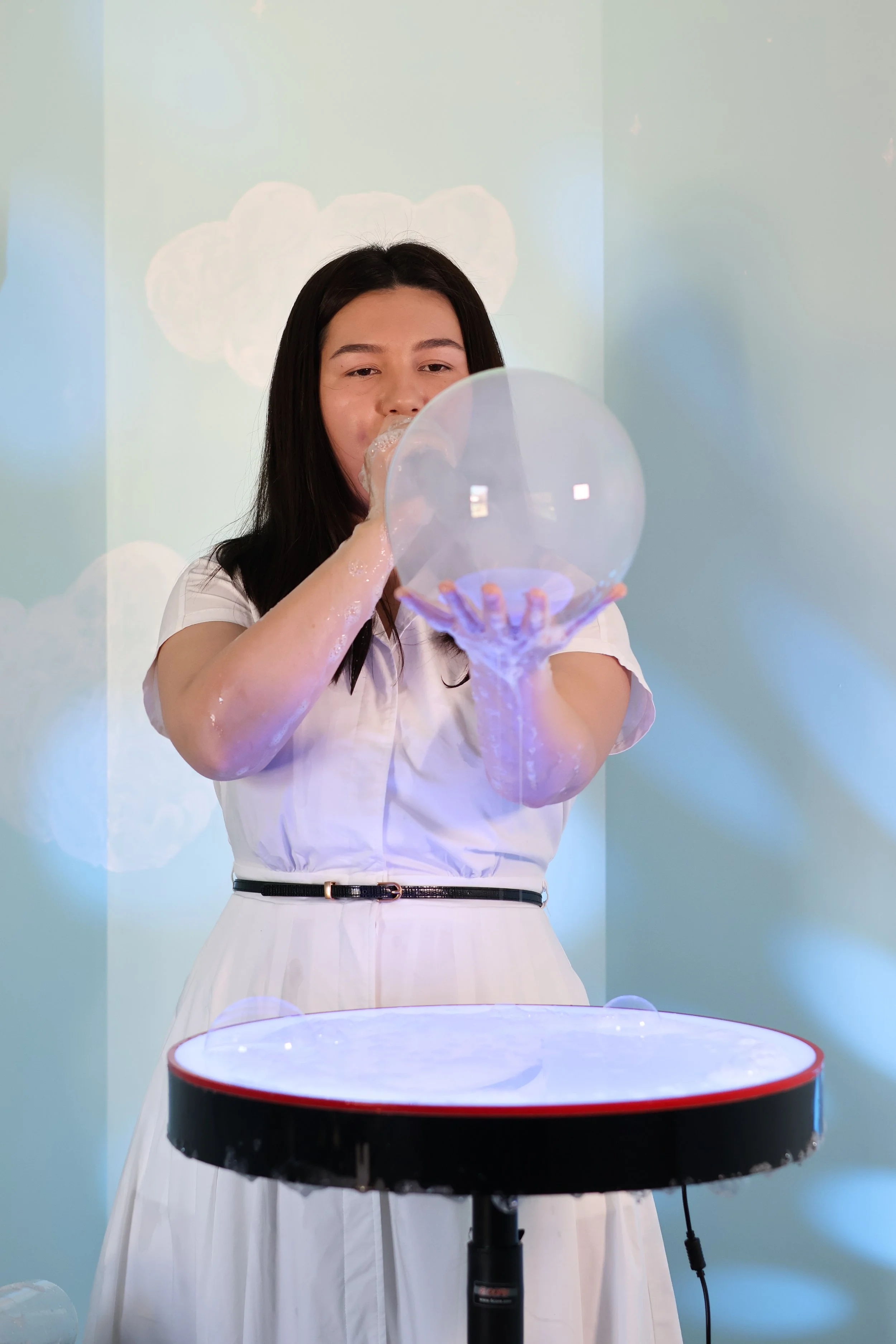 A woman in a white dress blowing bubbles through a large bubble wand, with a bubble-making machine on the table in front of her.