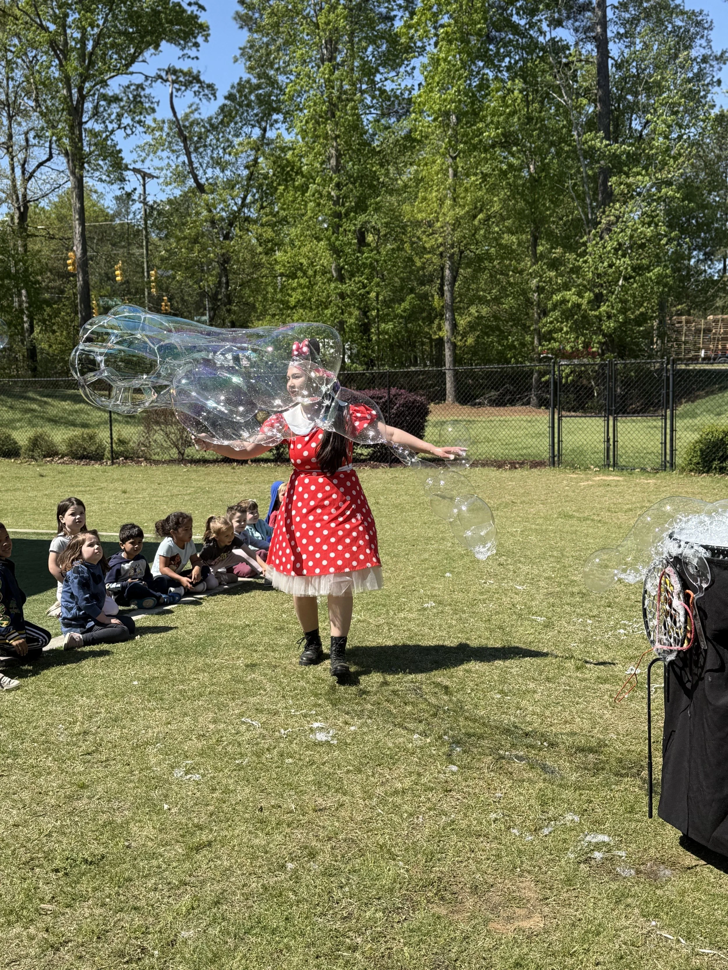 A young girl dressed in a red polka dot dress with a white collar and a pink bow headband is creating large soap bubbles outdoors on a sunny day. She is surrounded by children sitting on the grass, watching her. Trees with green leaves and a chain-li