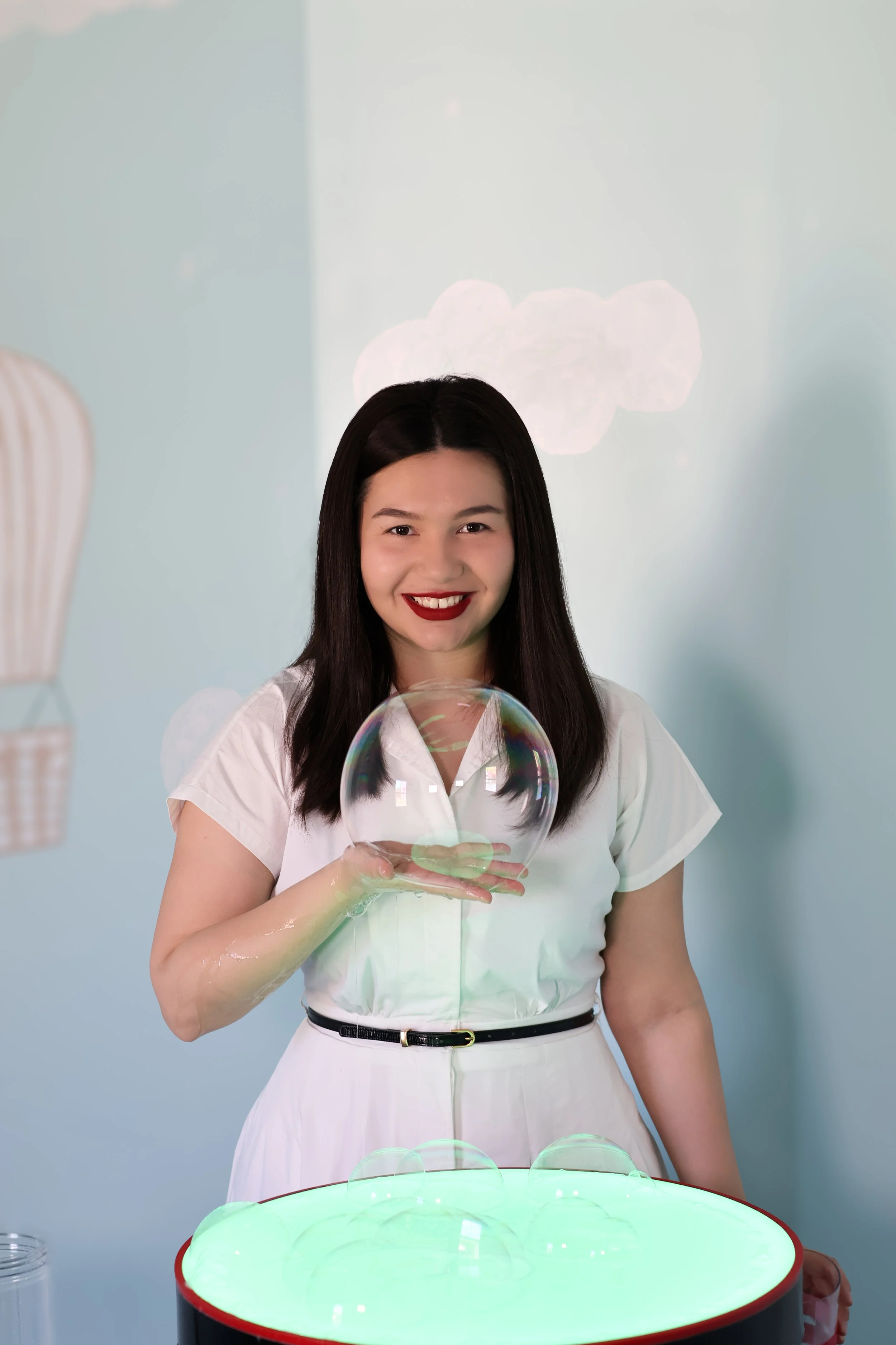 A woman with dark hair and red lipstick holding a large bubble in front of a light blue wall with cloud illustrations, standing over a bubble machine creating multiple bubbles.