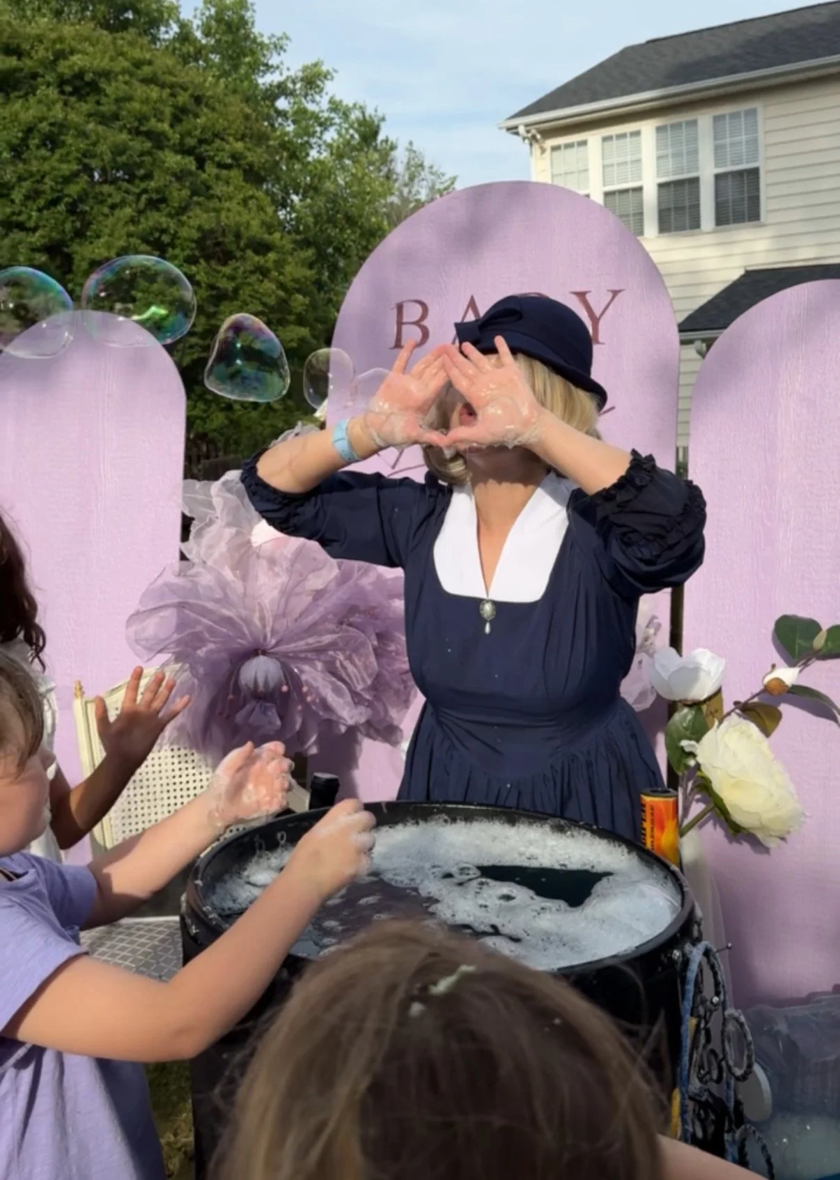 Woman dressed as a 1950s nanny making soap bubbles for children at an outdoor birthday party with a pink backdrop that says 'BABY' and large paper flowers.