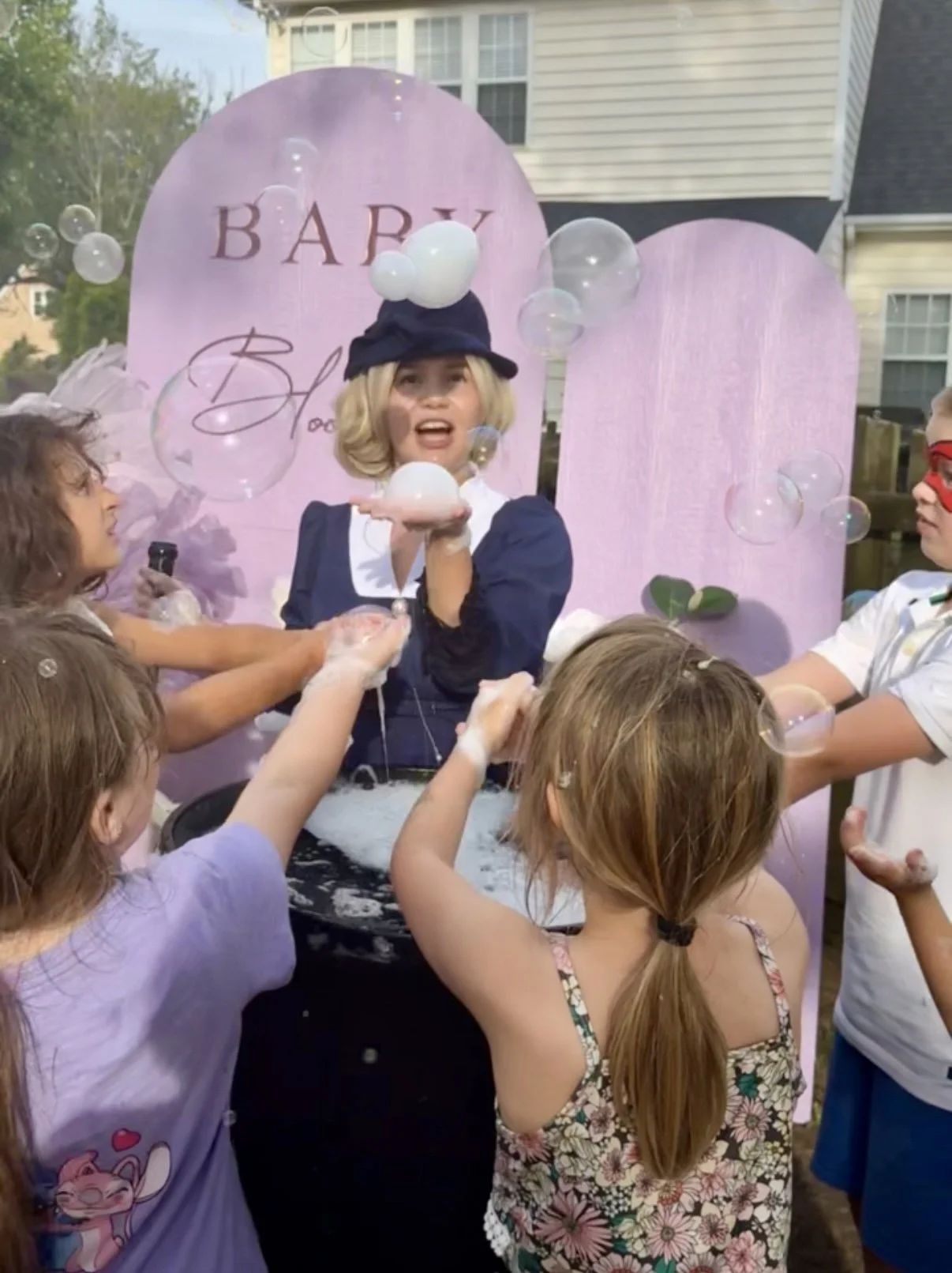 A woman dressed as a vintage doll, surrounded by children, is at a bubble-blowing station for kids' party.