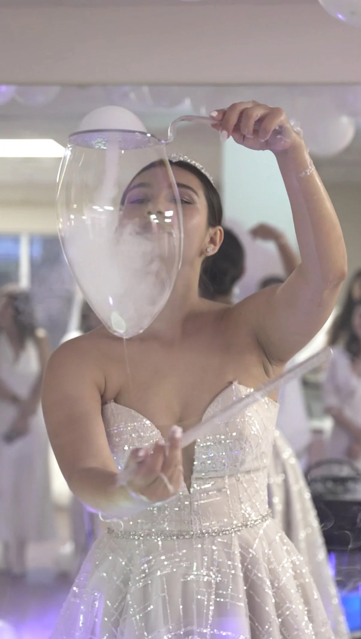 A young woman in a sparkly, strapless gown holding a large glass with shooting smoke or vapor, smiling at a festive event surrounded by other people.