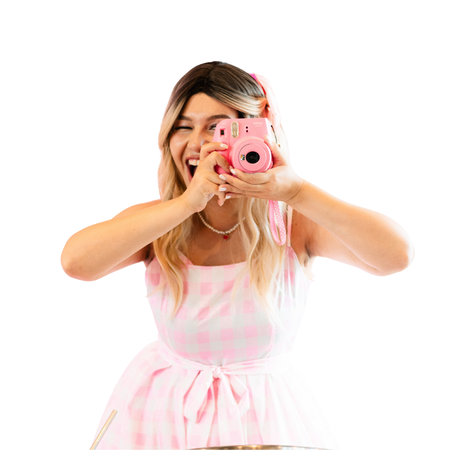 A young woman with blonde hair wearing a pink and white checkered dress, taking a picture with a pink instant camera, smiling and winking.