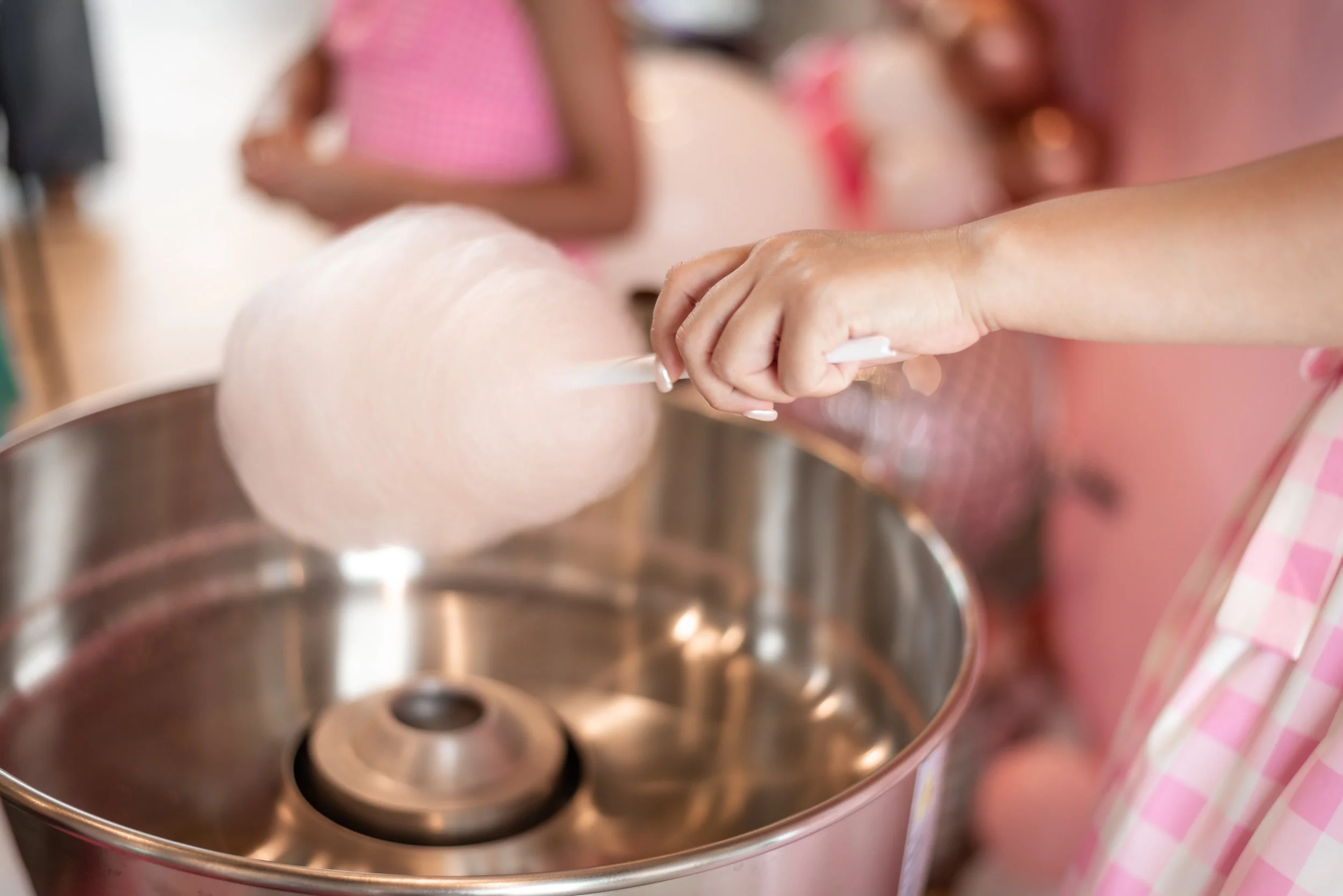 Person making cotton candy with a pink machine, with children in the background at a fair or carnival.