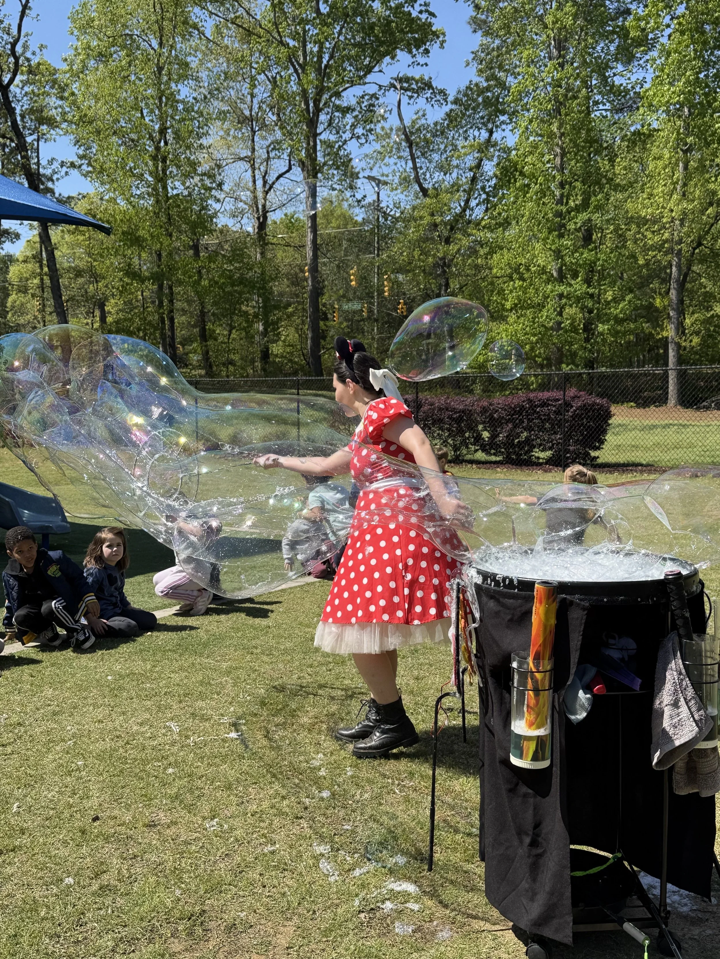 A woman dressed as Minnie Mouse is making large soap bubbles for children in a park on a sunny day, with trees and a fence in the background.