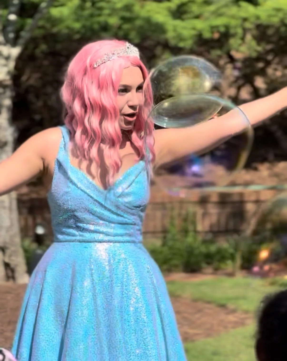 Woman with pink hair wearing a blue dress and silver tiara, holding large soap bubbles outdoors.