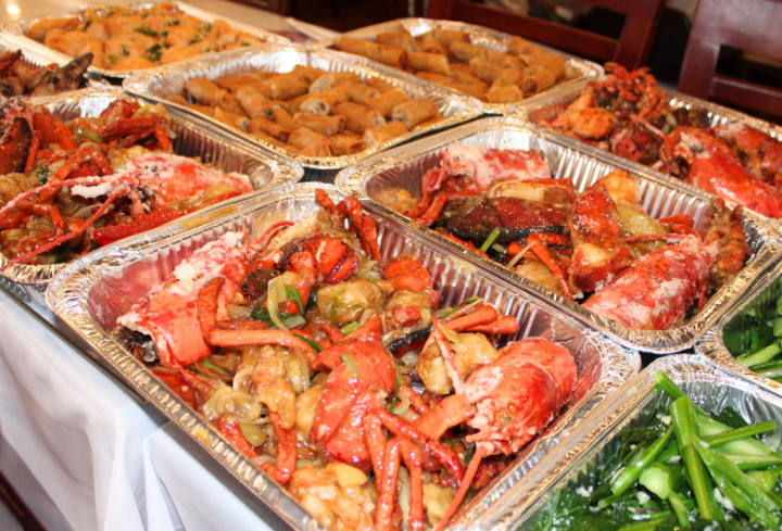 Large trays of cooked seafood dishes, including lobster, crab legs, and stir-fried vegetables, along with spring rolls and green vegetables, set on a buffet table.