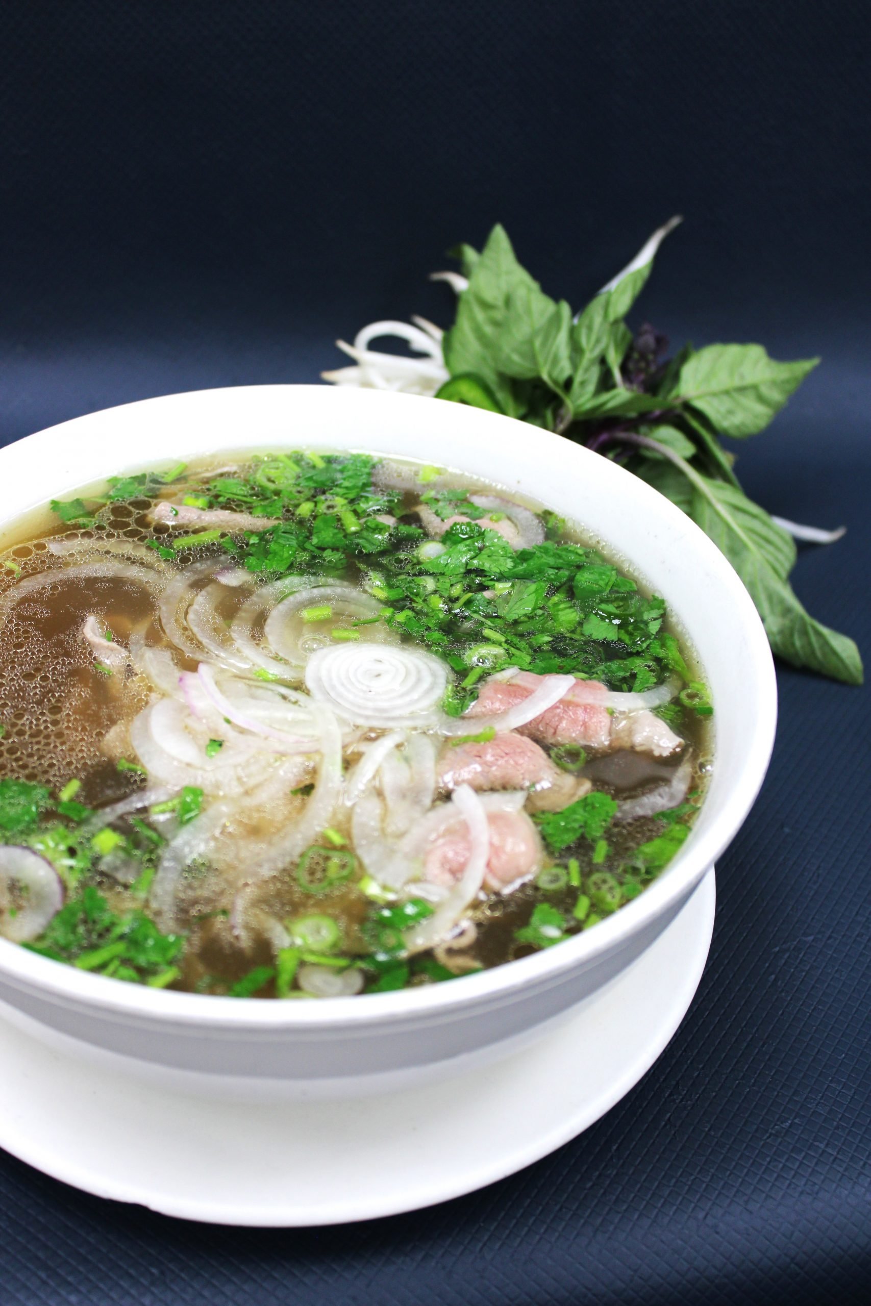 A bowl of Vietnamese pho with sliced onions, chopped green onions, herbs, and thin slices of meat, served on a white plate with fresh herbs in the background.