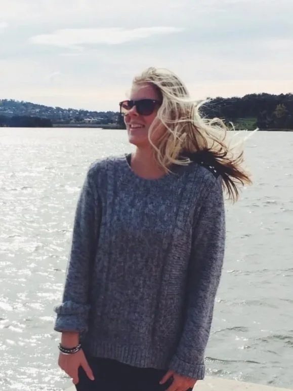 A woman with blonde hair wearing sunglasses and a gray sweater stands near a body of water with trees and hills in the background.