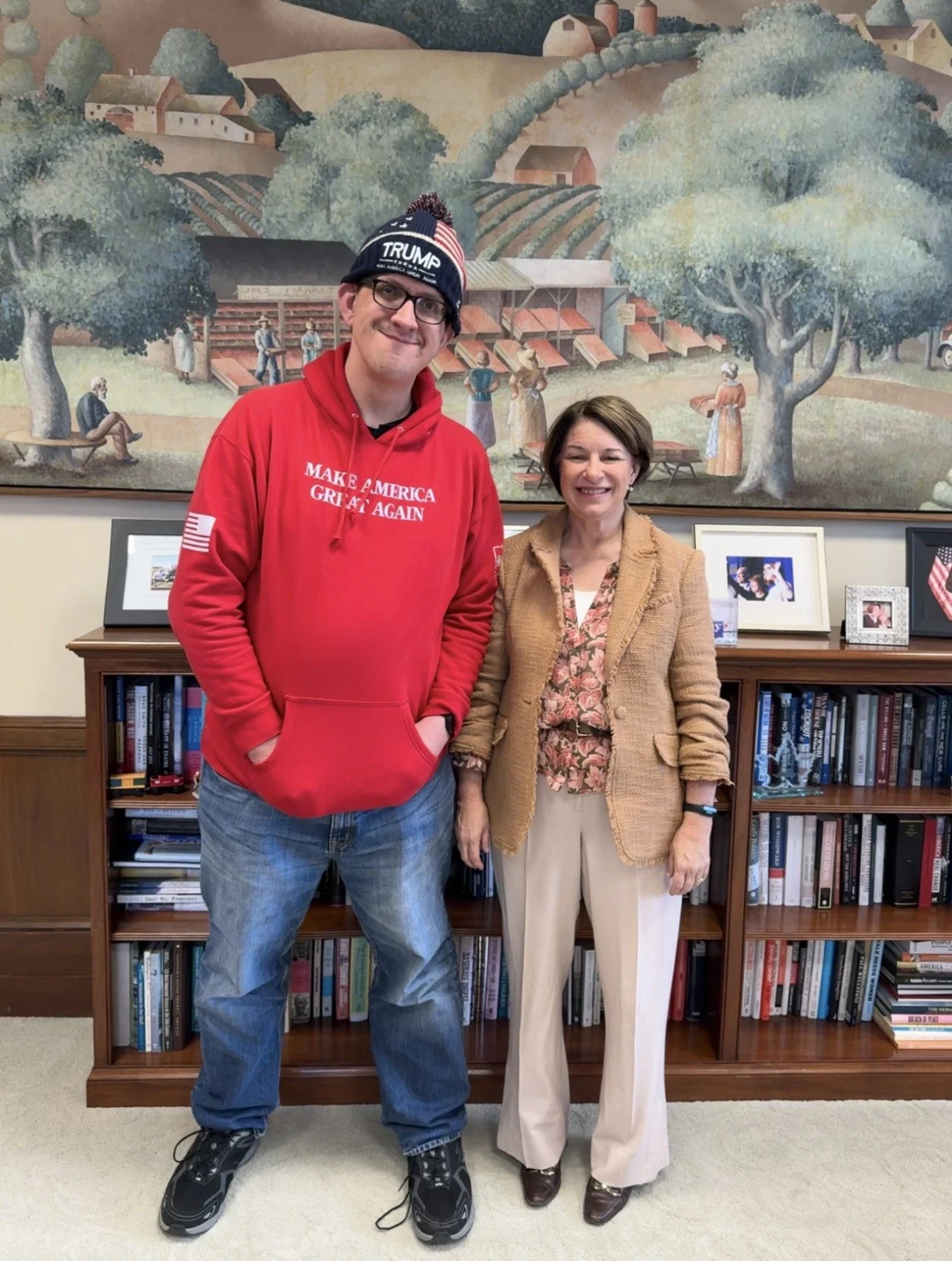 A man wearing a 'Make America Great Again' hoodie and a 'Trump' hat standing next to a woman in a beige blazer and patterned blouse, both smiling in front of a bookshelf and a large mural of a countryside scene.