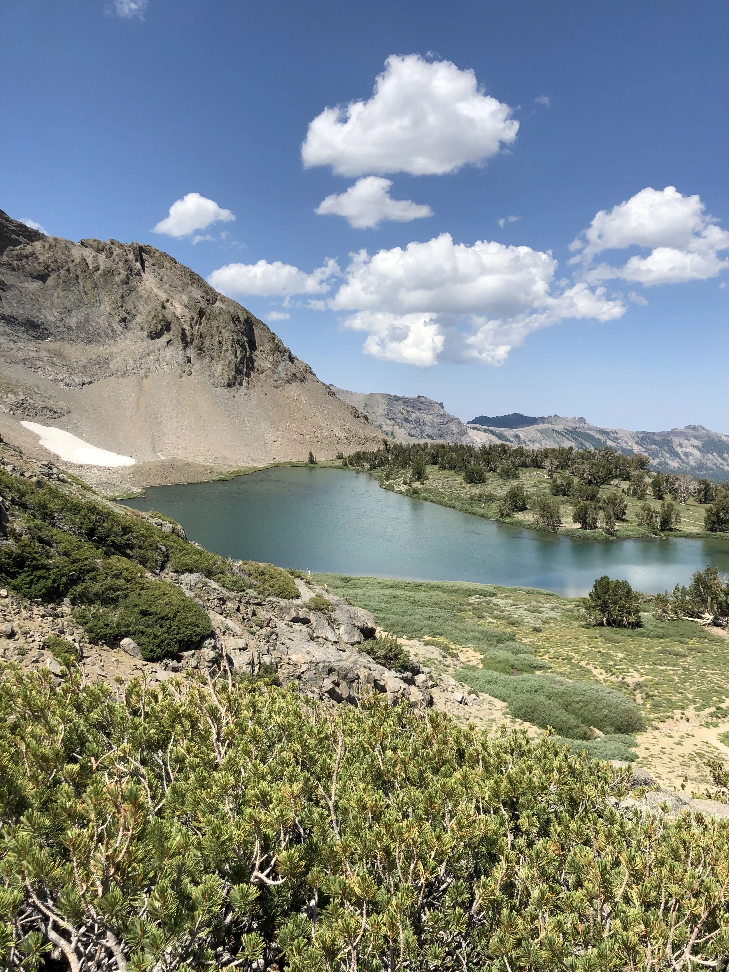 A scenic mountain landscape with a lake, rocky slopes, green vegetation, and a partly cloudy sky.