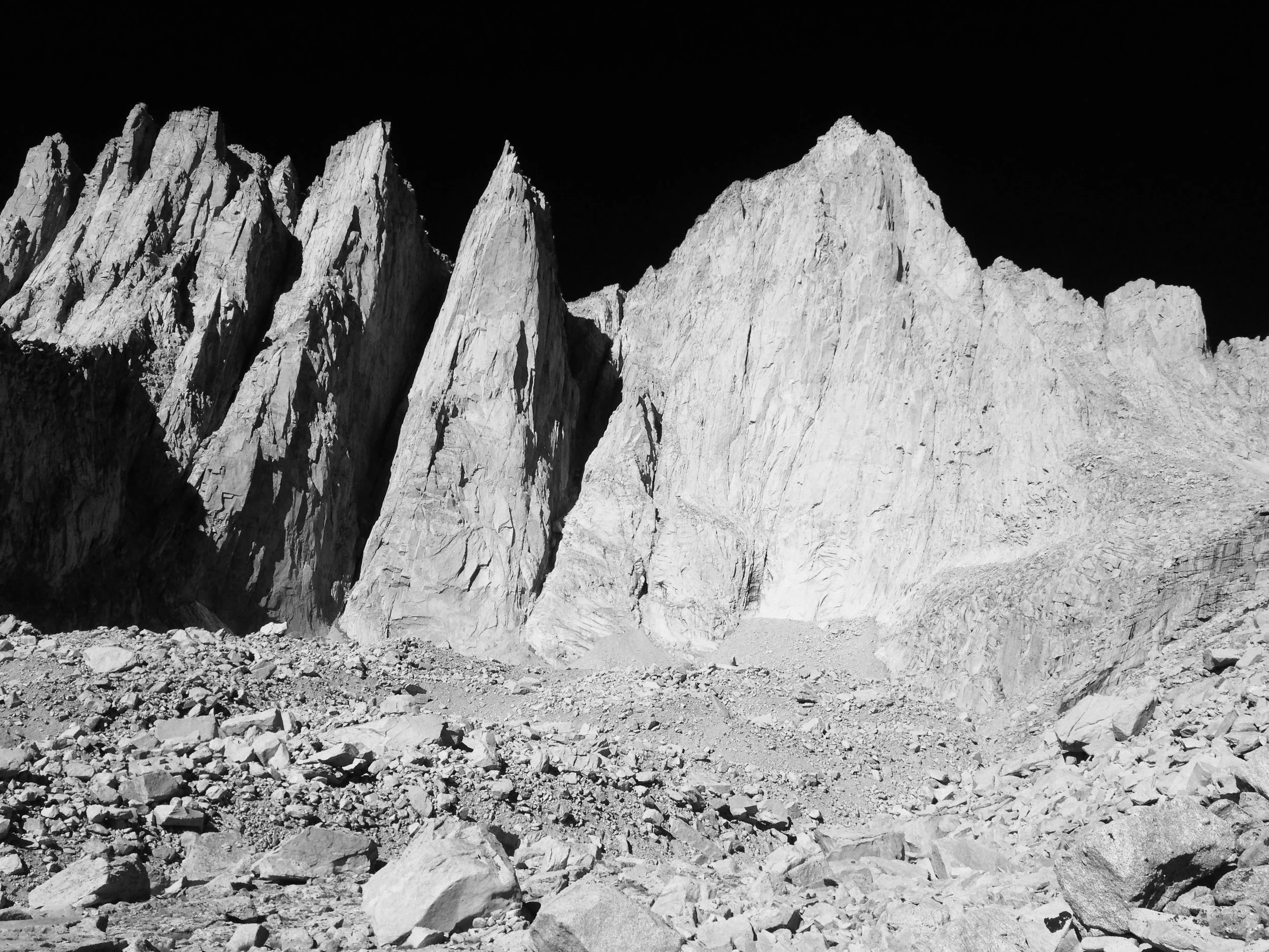 Black and white photograph of tall, jagged mountain peaks with a dark sky in the background and rocky terrain in the foreground.
