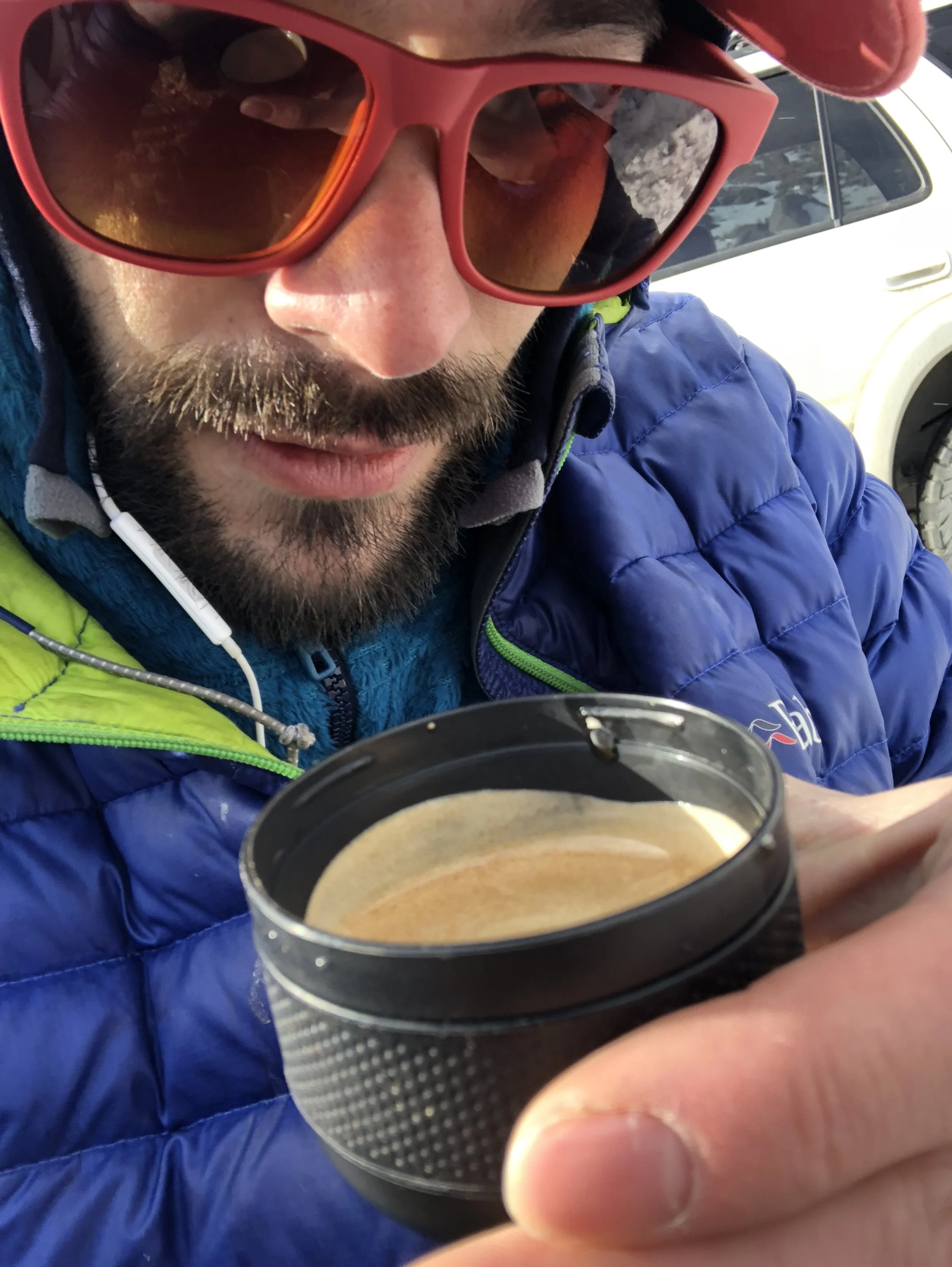 Close-up of a man in outdoor gear holding a cup of coffee, wearing red sunglasses and a blue jacket, with snow-covered mountains in the background.