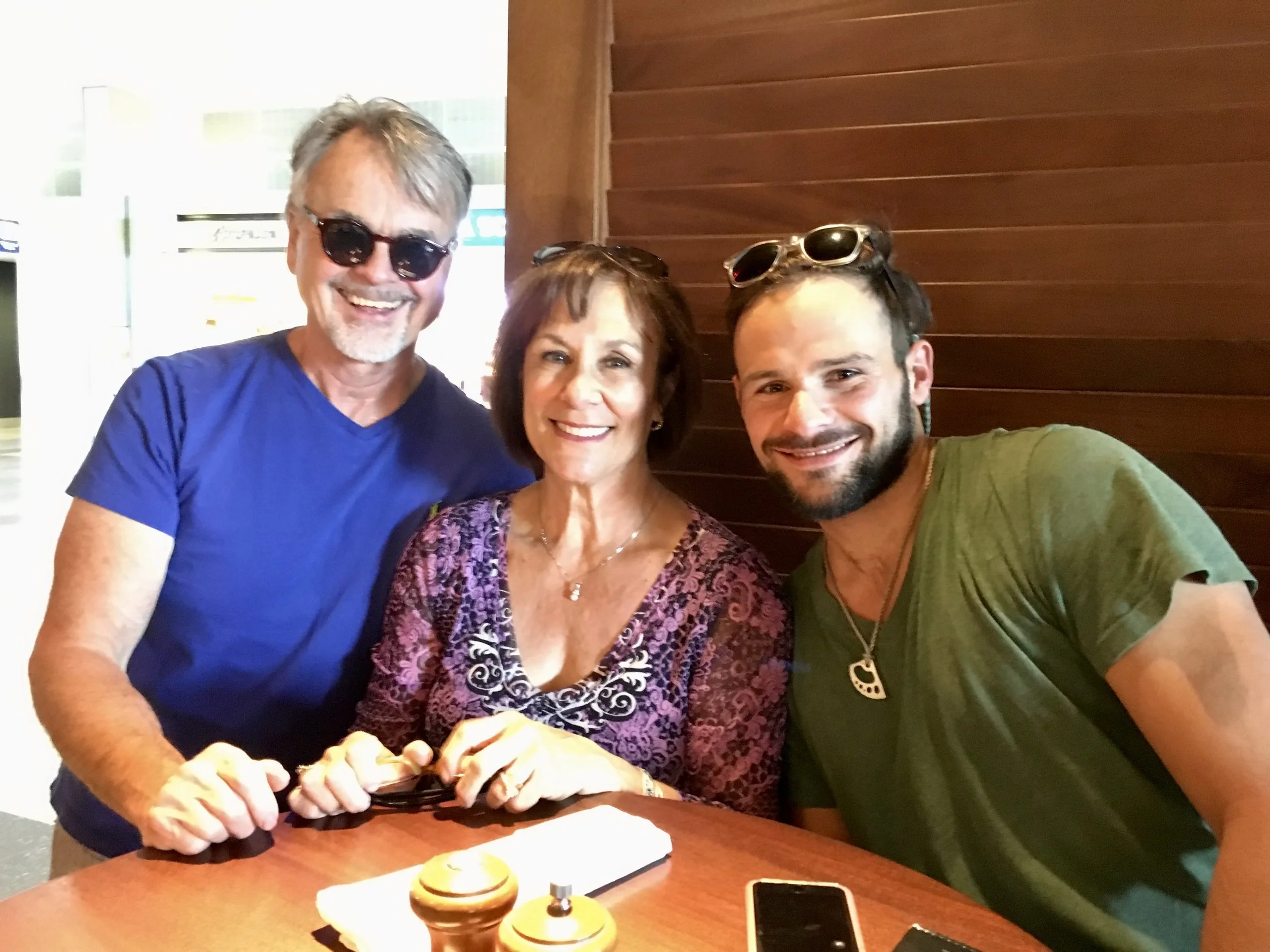Three smiling people sitting at a table in a restaurant. An older man wearing sunglasses in a blue t-shirt, a middle-aged woman with dark brown hair in a floral purple top, and a young man with sunglasses on his head in a green t-shirt.