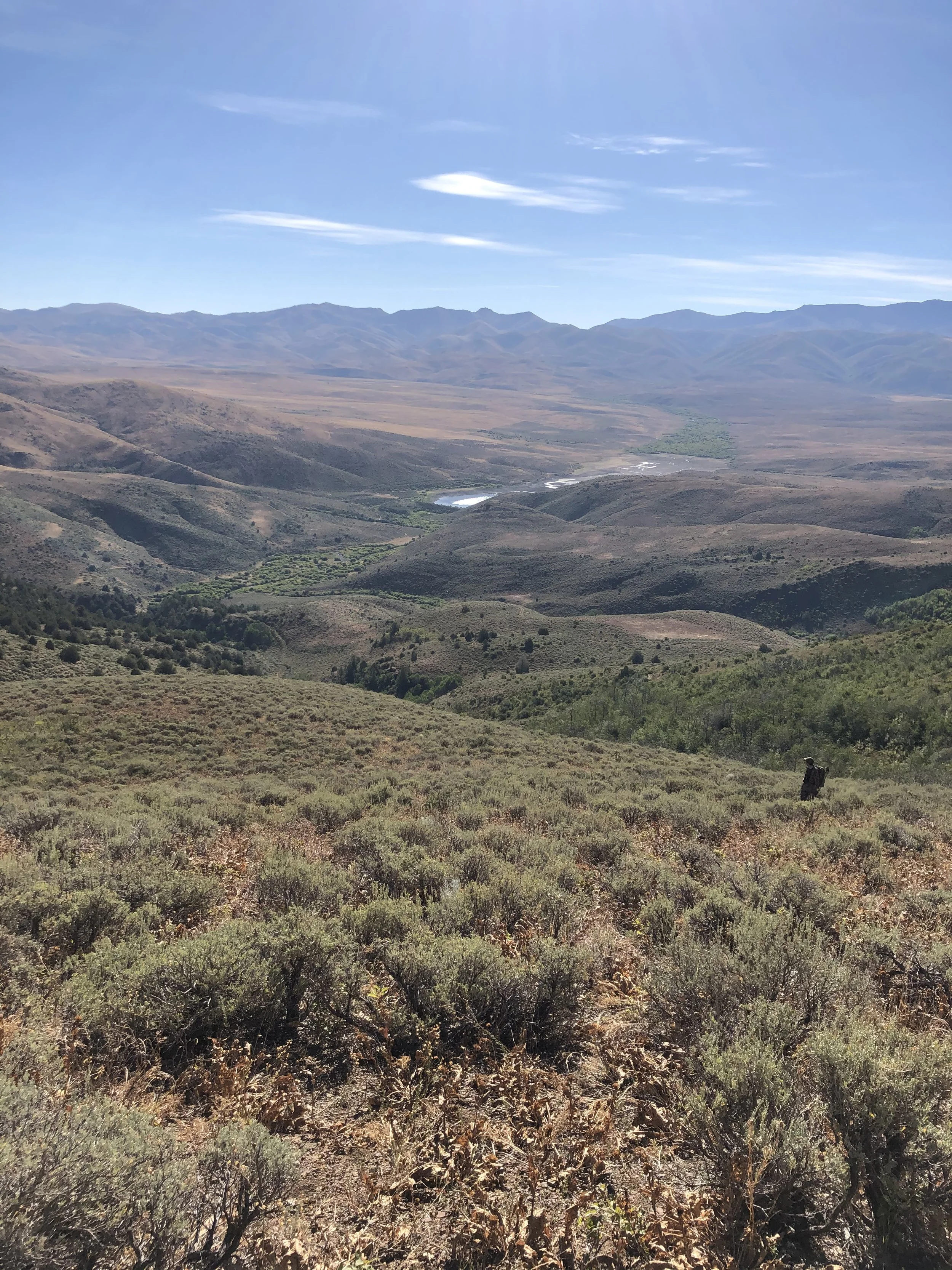 Mountain landscape with rolling hills, sparse vegetation, distant mountains, blue sky, and some clouds.