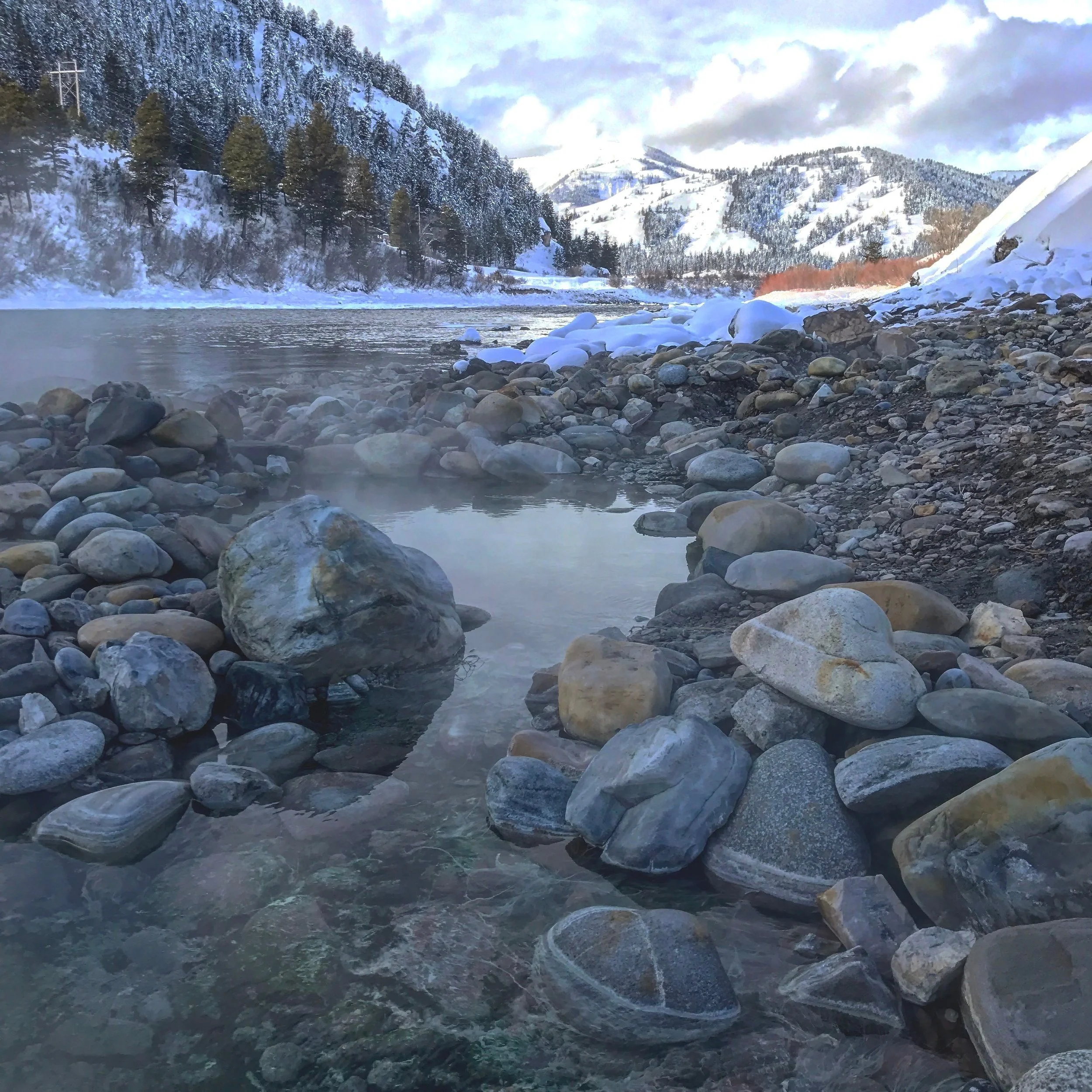 A rocky riverbank with snow and ice, mountains covered in snow, and a partly cloudy sky in the background.