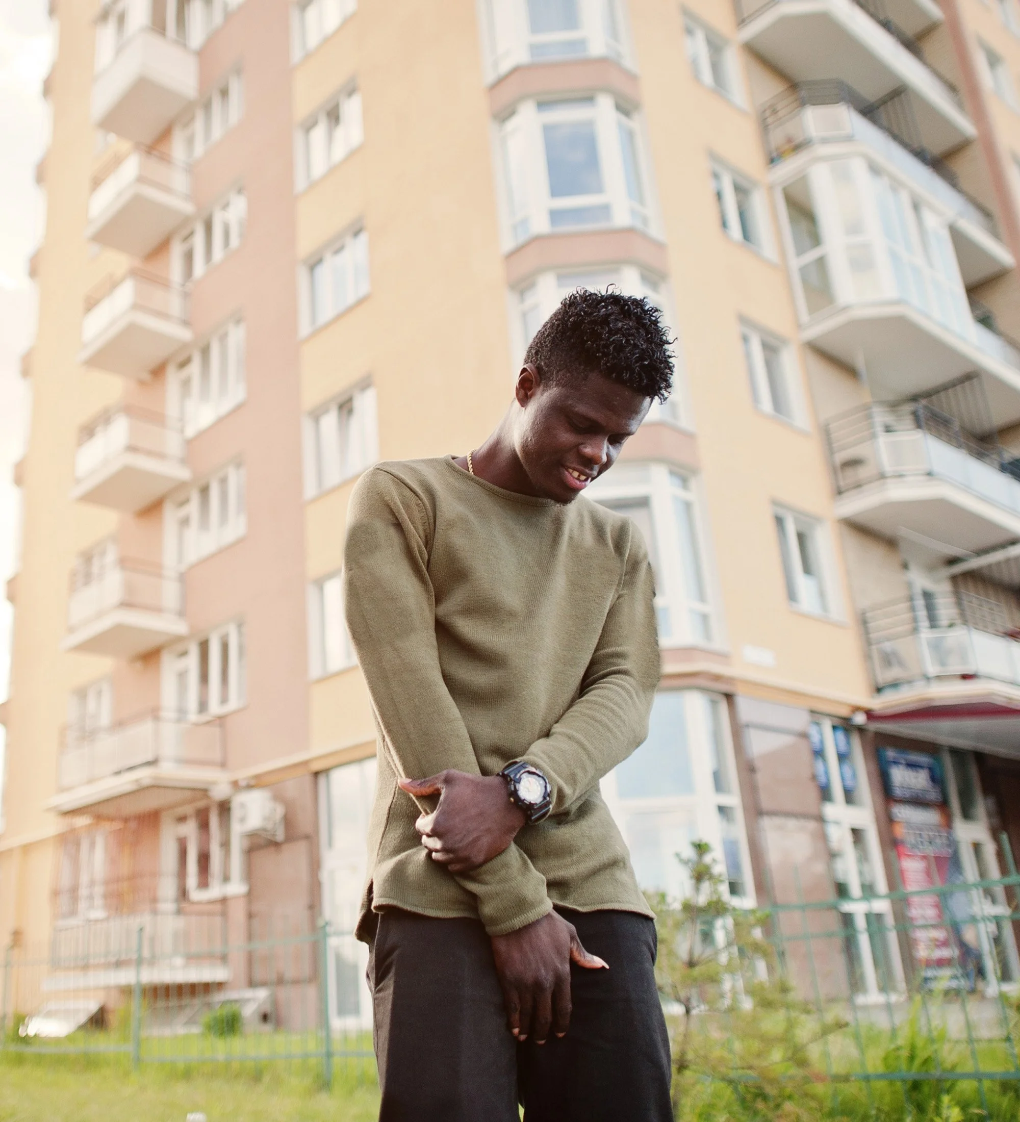 A young man with curly hair wearing a green long-sleeve shirt and black pants stands outdoors in front of a multi-story apartment building. He appears to be in discomfort, holding his side with one hand and touching his stomach with the other.