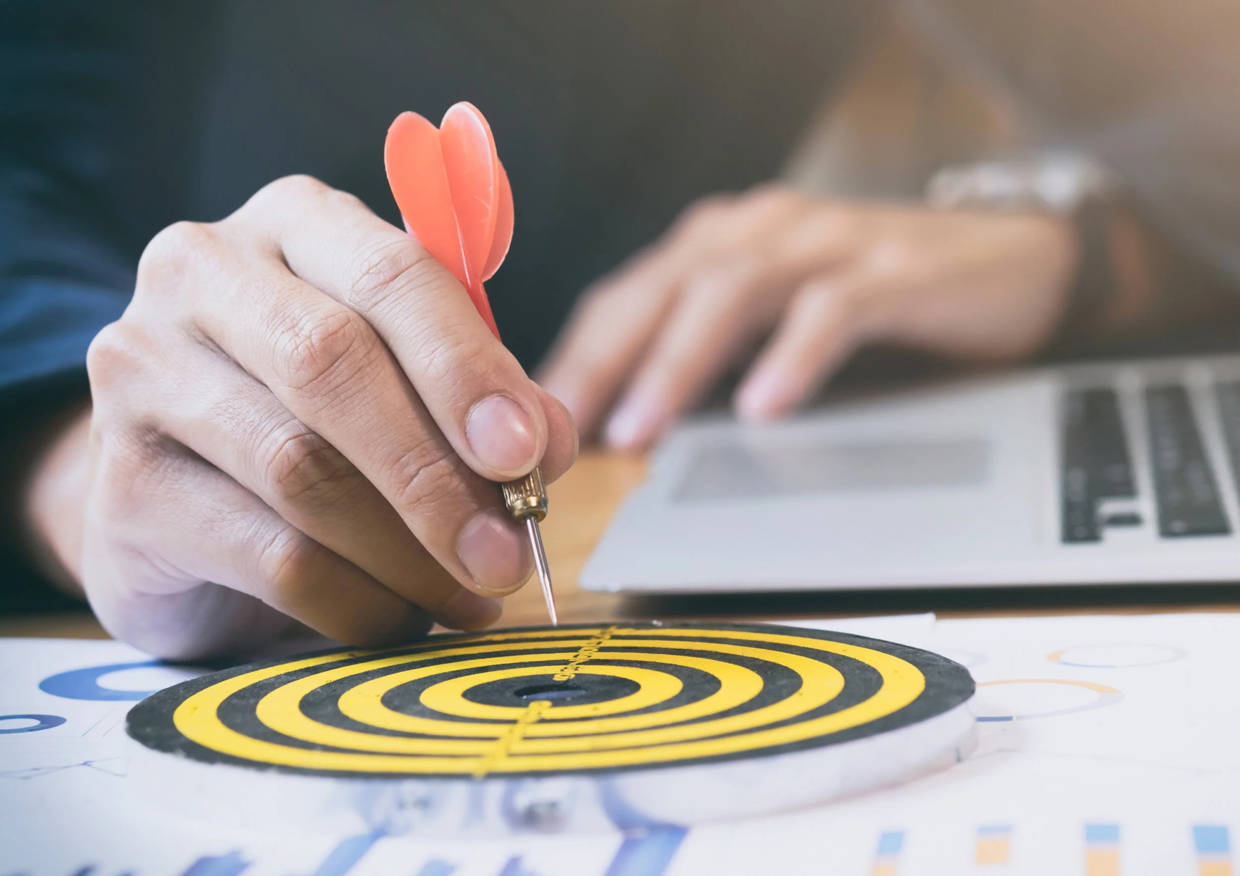 A person is using a dart with orange fins to aim at the center of a black and yellow target on a table, with a laptop in the background.
