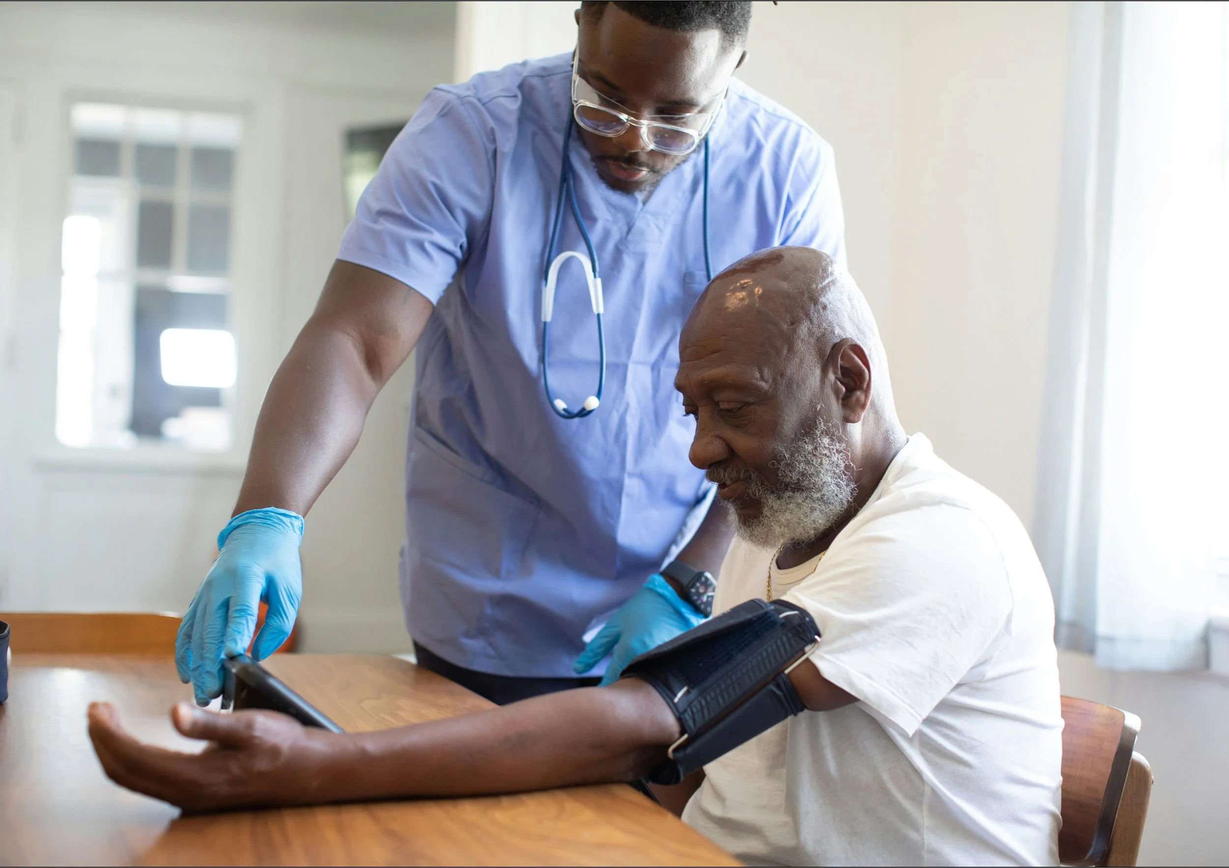 A healthcare professional in scrubs and gloves measuring an elderly man's blood pressure using a cuff, in a bright room.