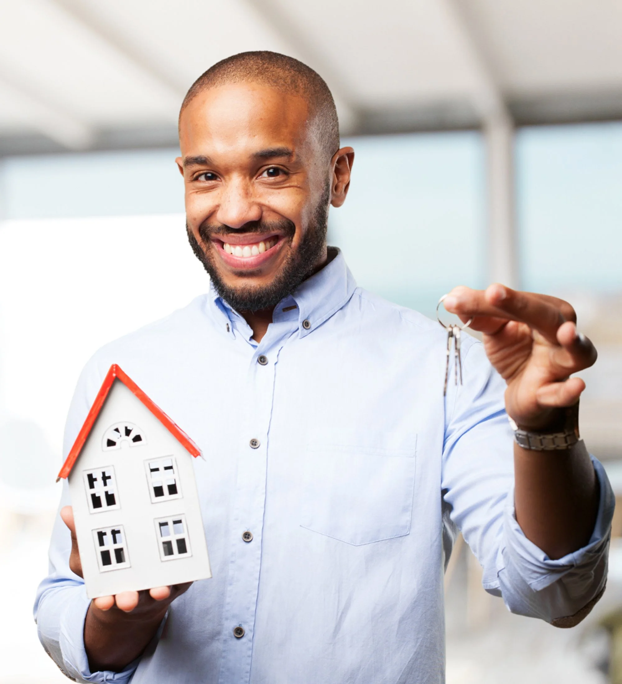 A man smiling and holding a small model house in one hand and a key in the other, in an indoor setting.