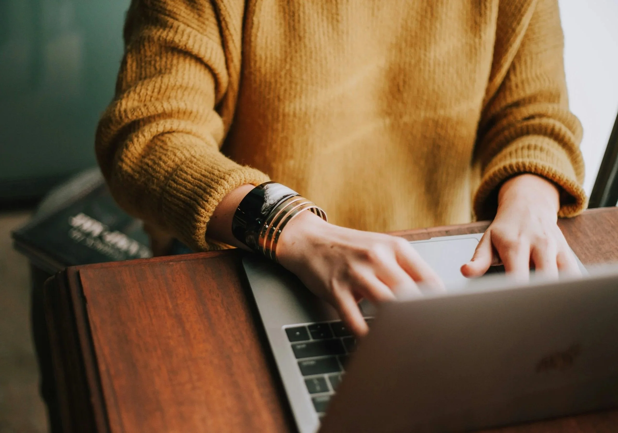 Person wearing a mustard-colored sweater typing on a laptop at a wooden desk.