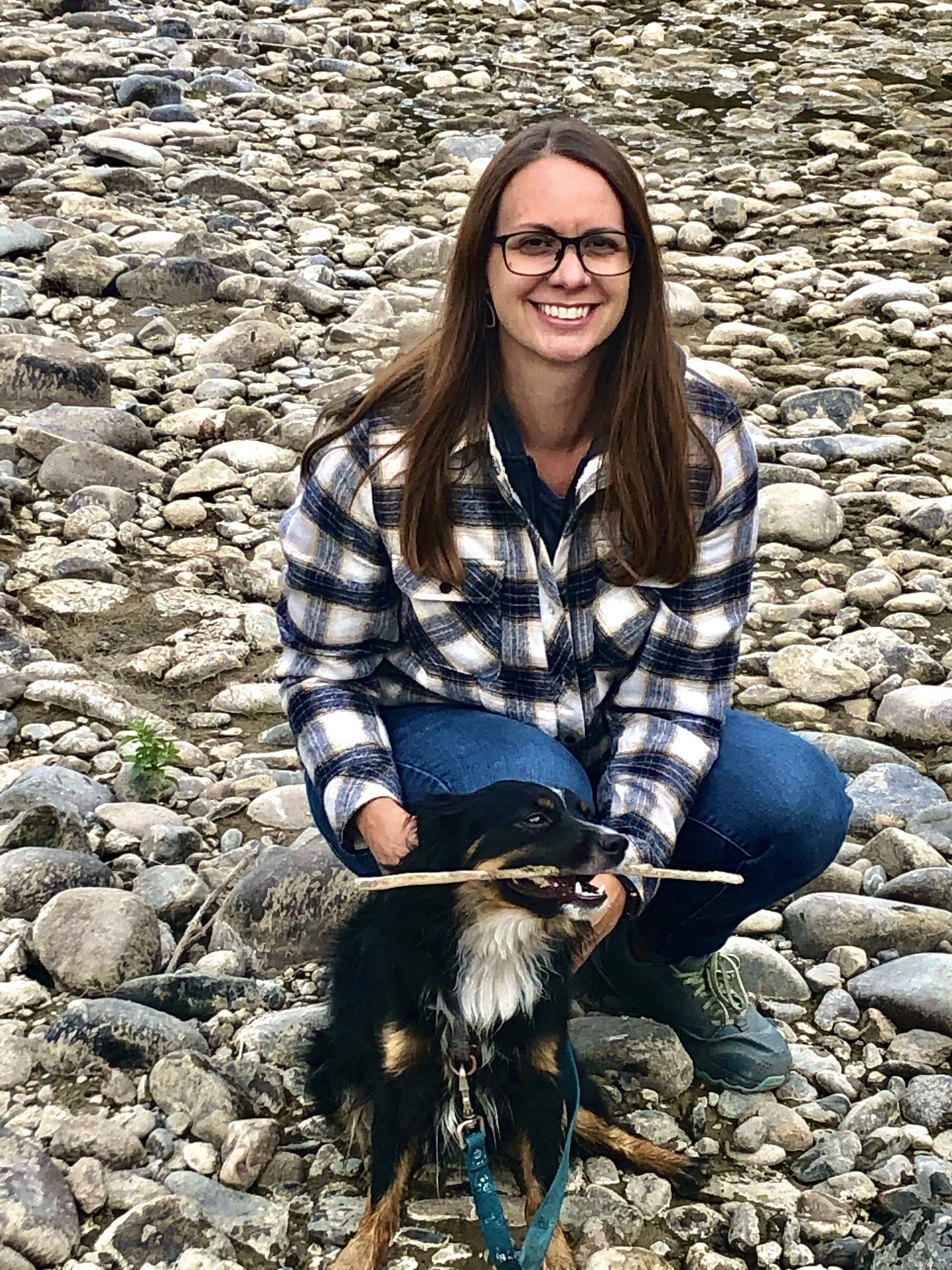 A woman with long brown hair and glasses, wearing a plaid shirt and jeans, crouching on a rocky riverbank with a black and tan dog, holding a stick in its mouth.