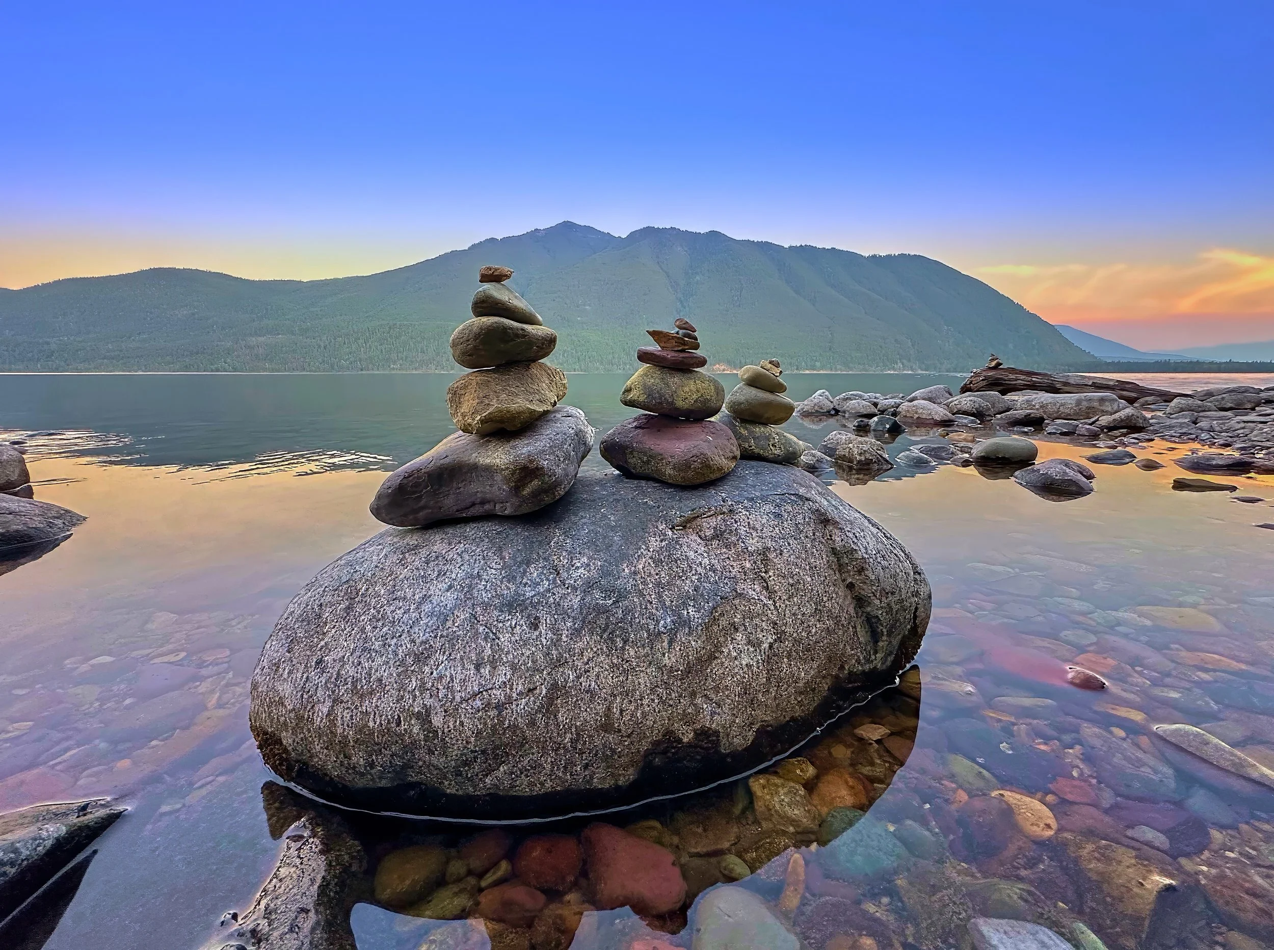 A large rock in the water with smaller stacked rocks on top, set against a mountain and colorful sky at sunset or sunrise.