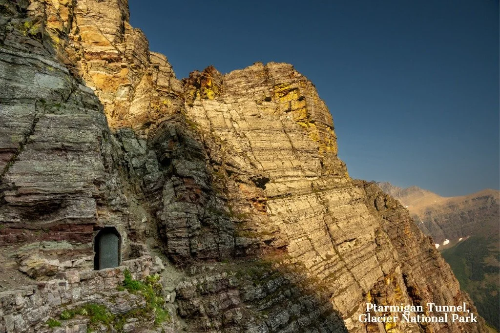 A rugged mountain cliff face with a tunnel entrance carved into the rock at the bottom left. The cliff has layers of sedimentary rock with some patches of green vegetation. The sky is clear and blue. Text in the bottom right corner reads "Ptarmigan Tunnel, Glacier National Park."