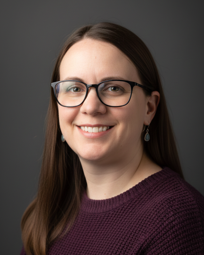 A woman  (Tracie ) with long brown hair, glasses, and earrings, smiling at the camera against a dark gray background.