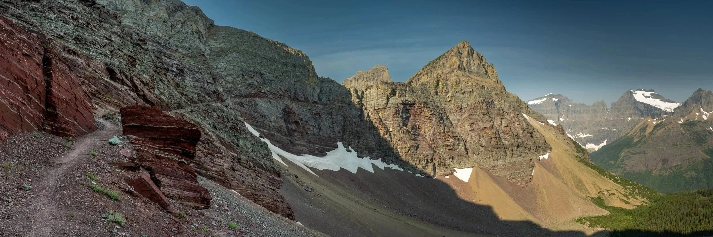 A rugged mountain trail winds through steep, colorful rock formations with snow patches, tall mountain peaks, and a green forested valley below under a clear sky. Ptarmigan Tunnel in Glacier National Park is visible