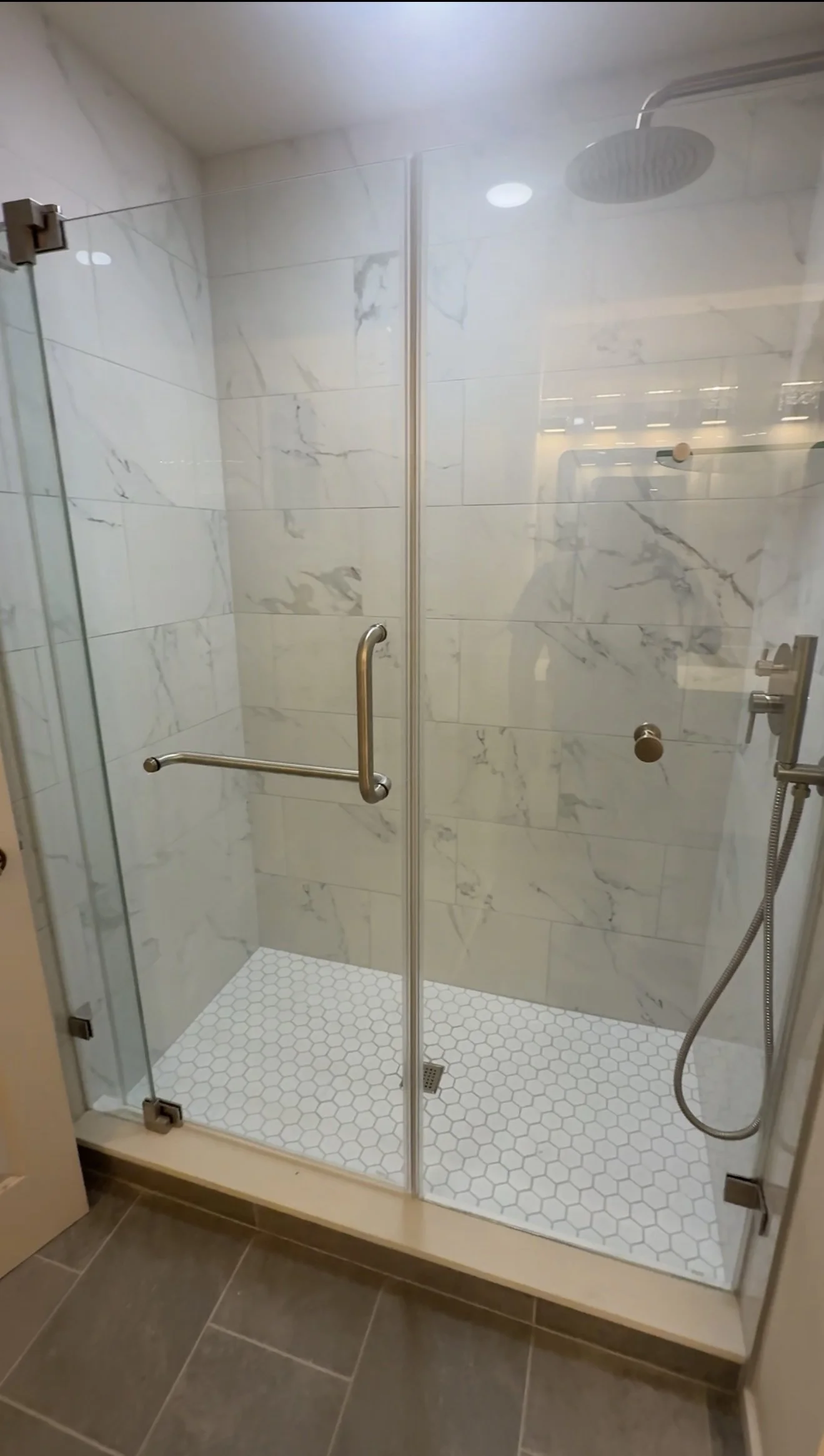 A modern glass-enclosed shower with white marble walls, a rain shower head, and a handheld shower. The shower has a sliding door with a metal handle, and the floor is covered with white hexagon tiles.