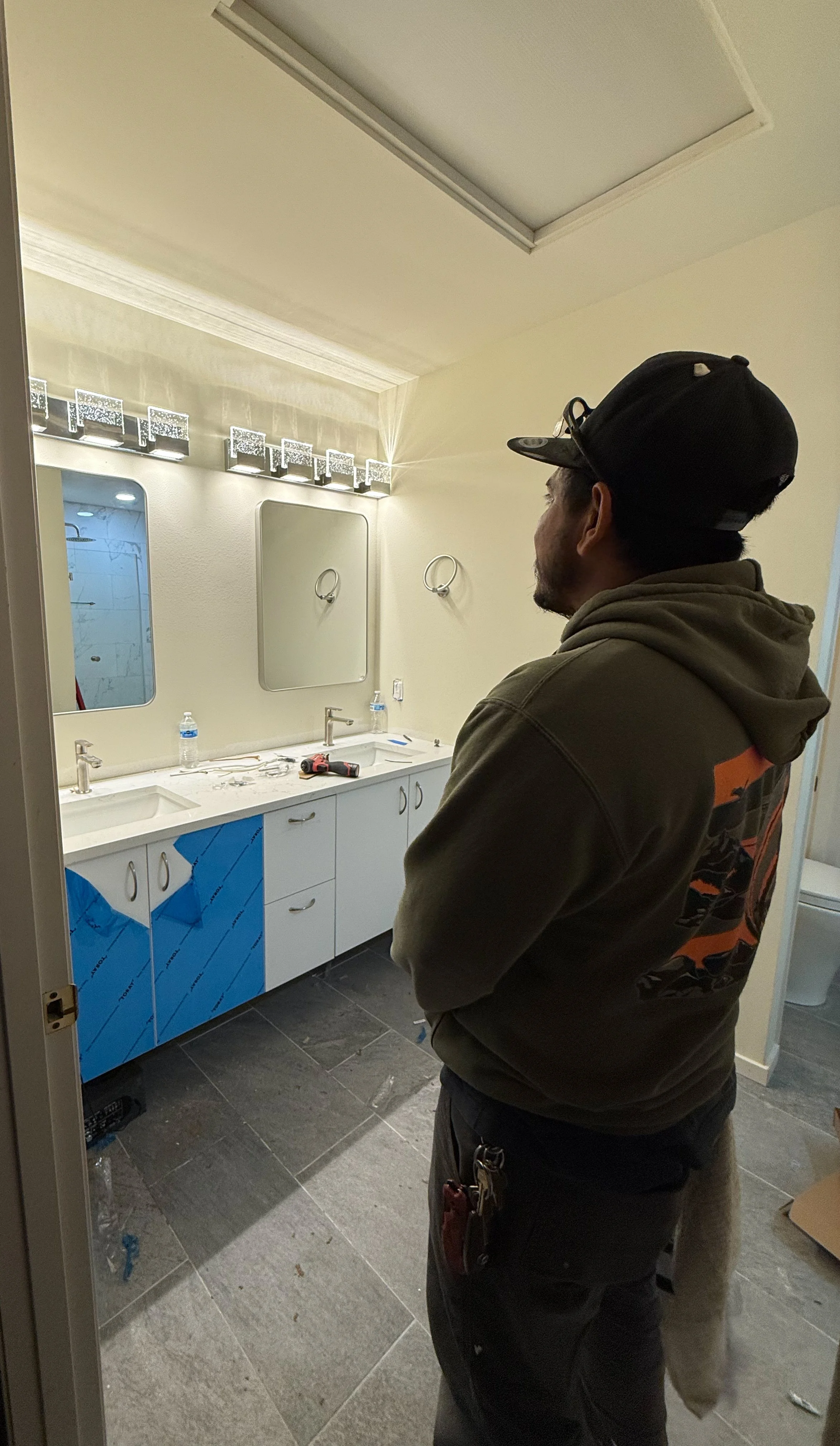 A man in a hoodie and cap standing in a bathroom under renovation, with cabinets covered in blue protective film, tools on the counter, and water bottles on the sink.
