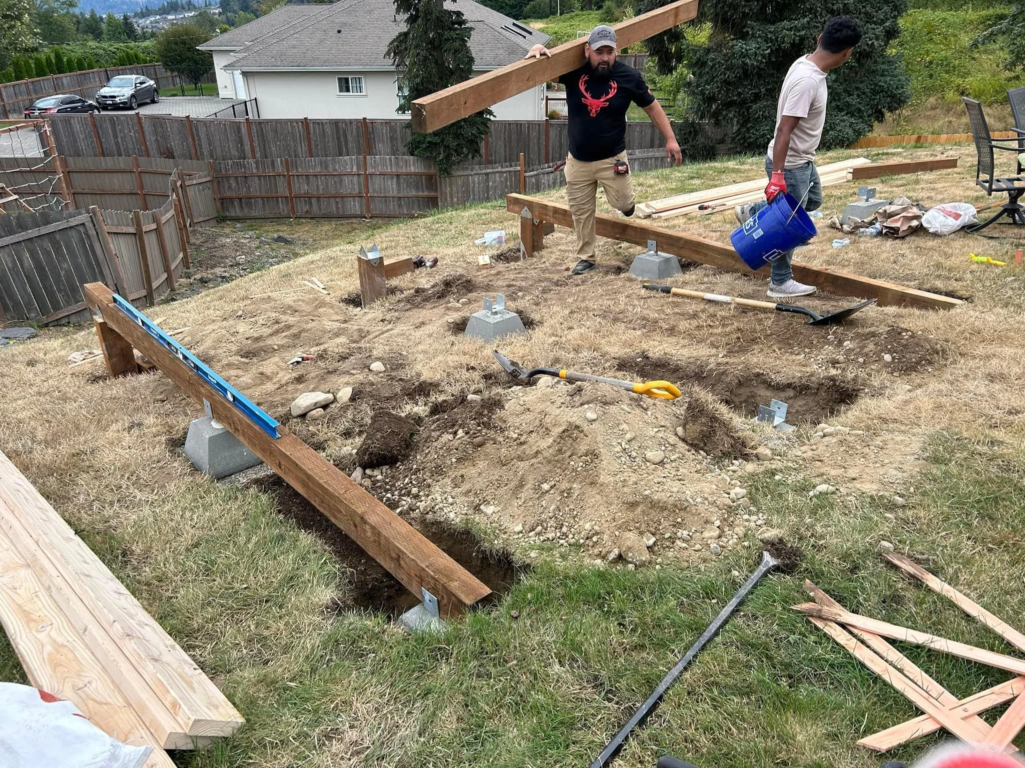 Two people working on a backyard construction project, setting wooden beams on concrete supports, with tools and materials scattered around.