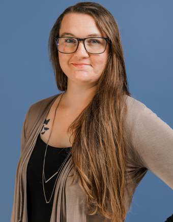 A woman with long brown hair, wearing glasses, a black top, and a beige cardigan, posing against a blue background.