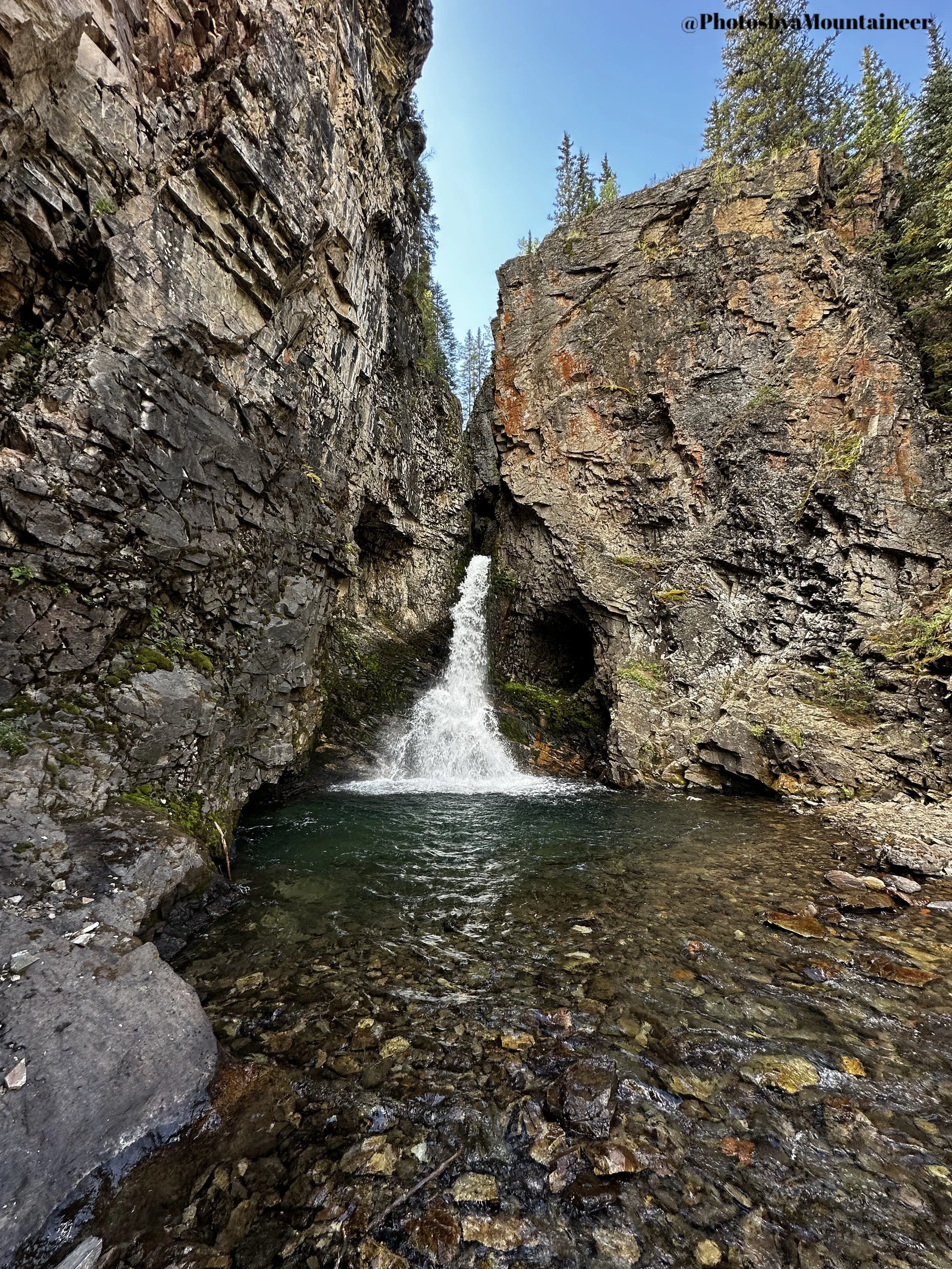 A small waterfall flowing into a rocky stream between two large steep cliffs, with pine trees and blue sky visible at the top of the cliffs.