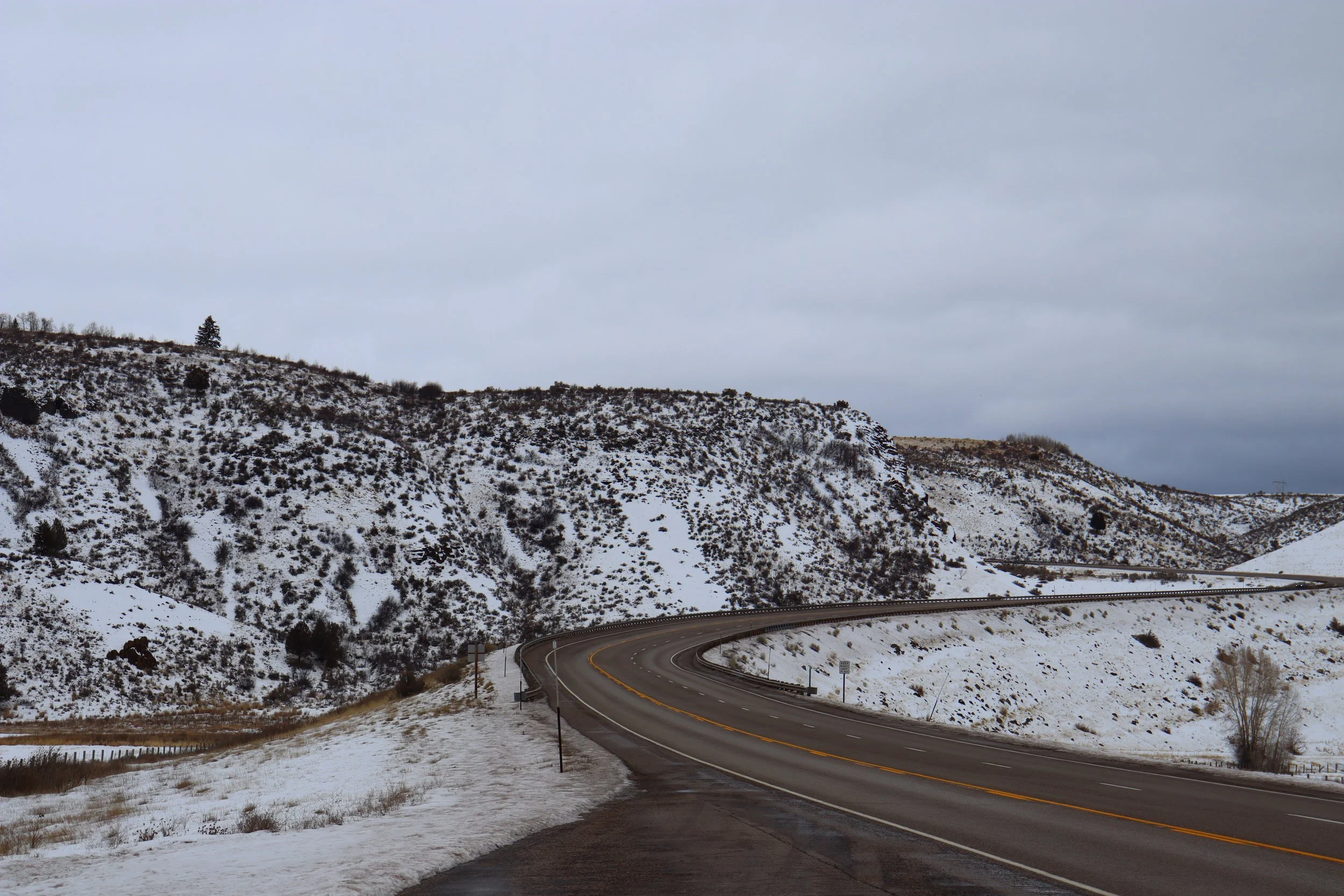 A winding mountain road with snow-covered slopes on either side, under an overcast sky.