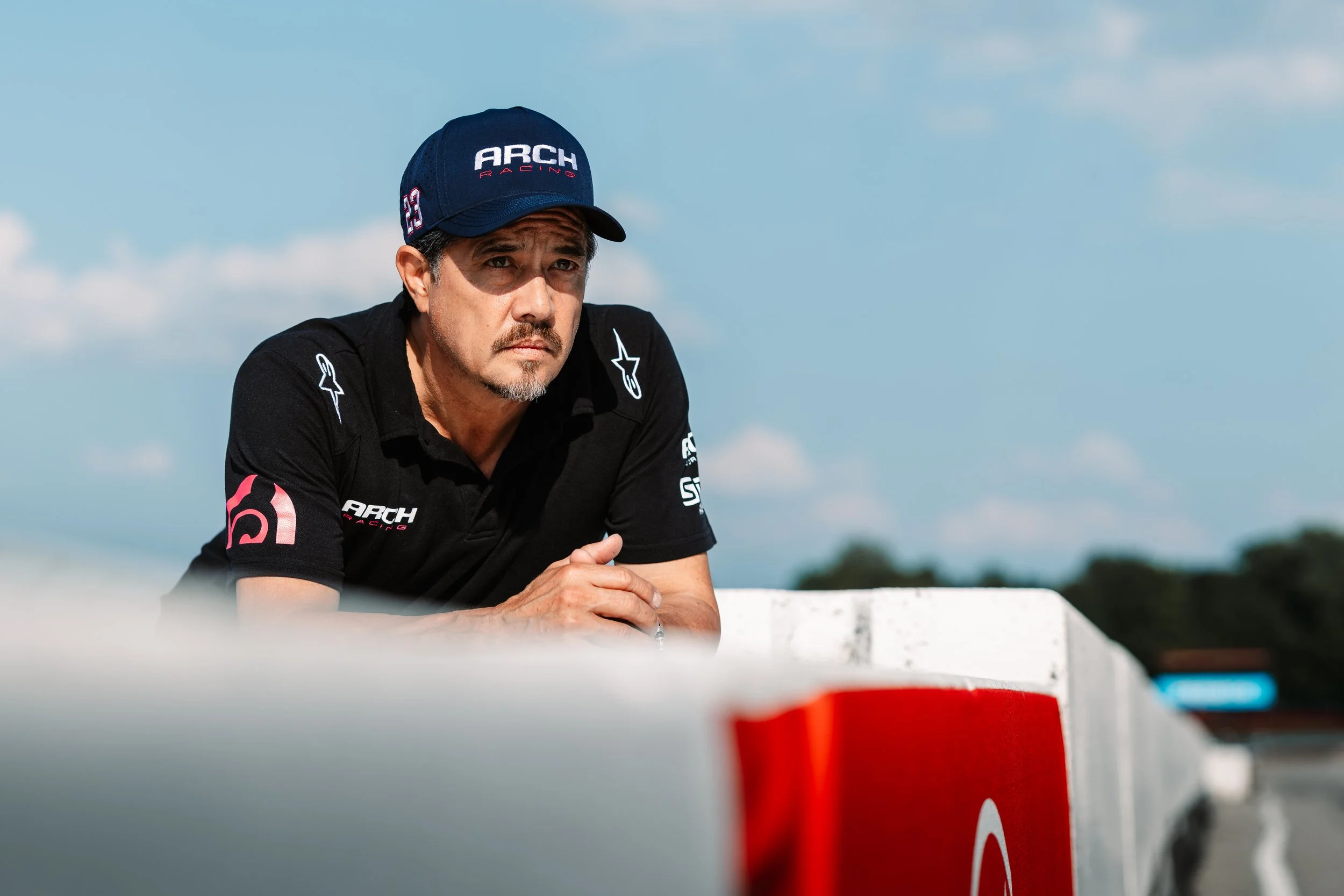 A man wearing a black polo shirt and a blue Cap with 'ARCH RACING' written on it, leaning on a white barrier with a serious expression, against a background of blue sky and clouds.