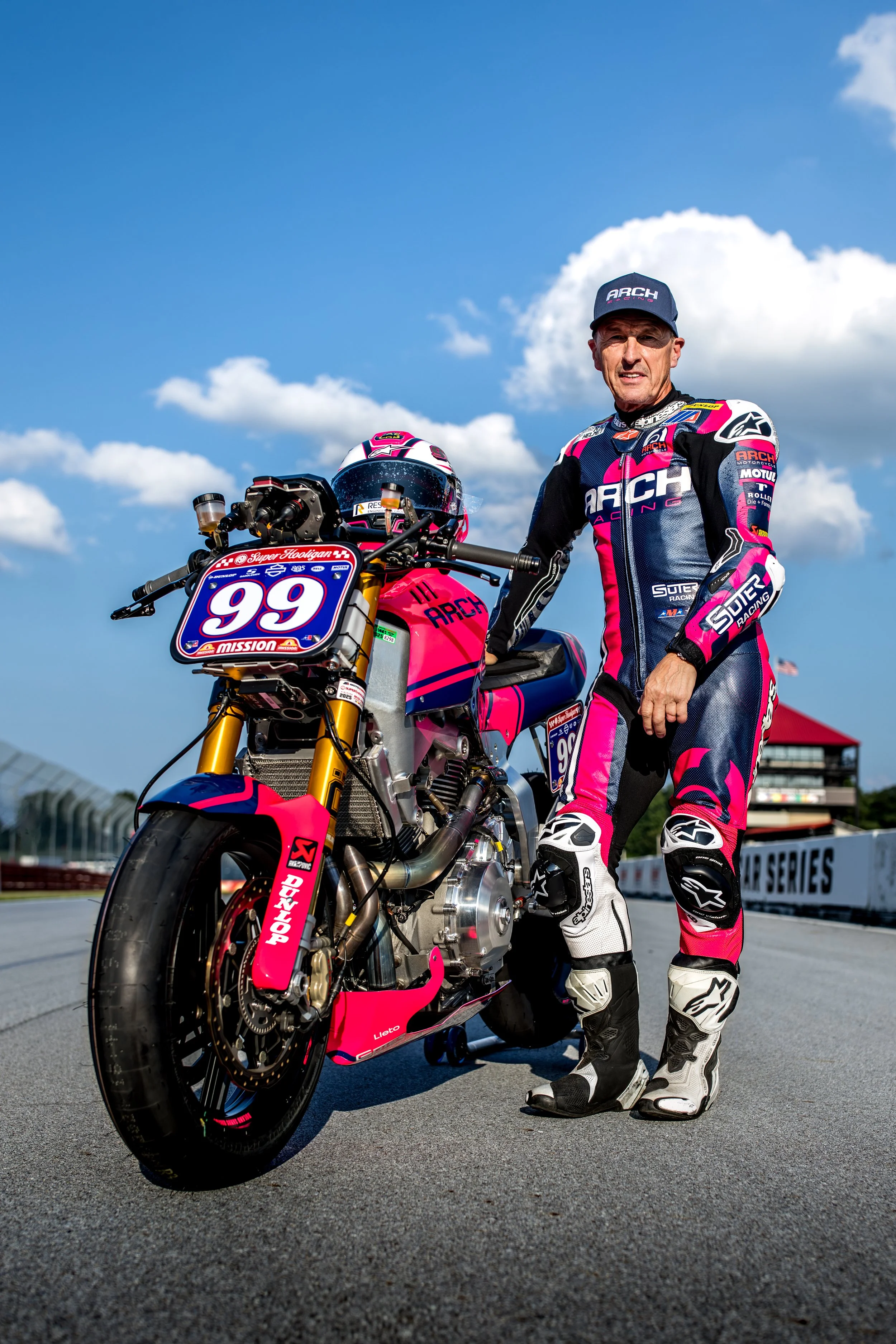 A race car driver in a pink, black, and white racing suit and hat standing next to a pink and black motorcycle on a race track under a blue sky with clouds.