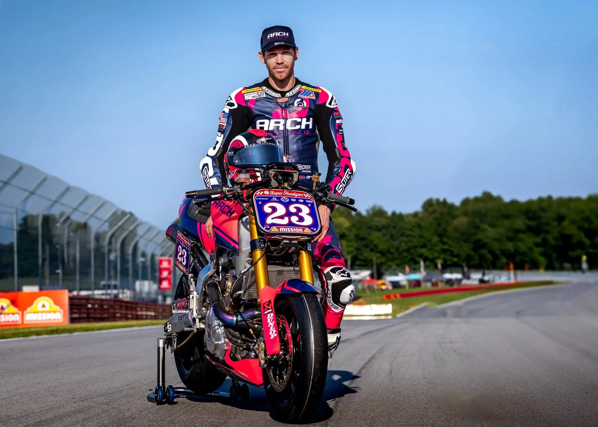A professional motorcycle racer standing on a race track with his motorcycle, wearing racing gear and a cap, with a blue sky and green trees in the background.