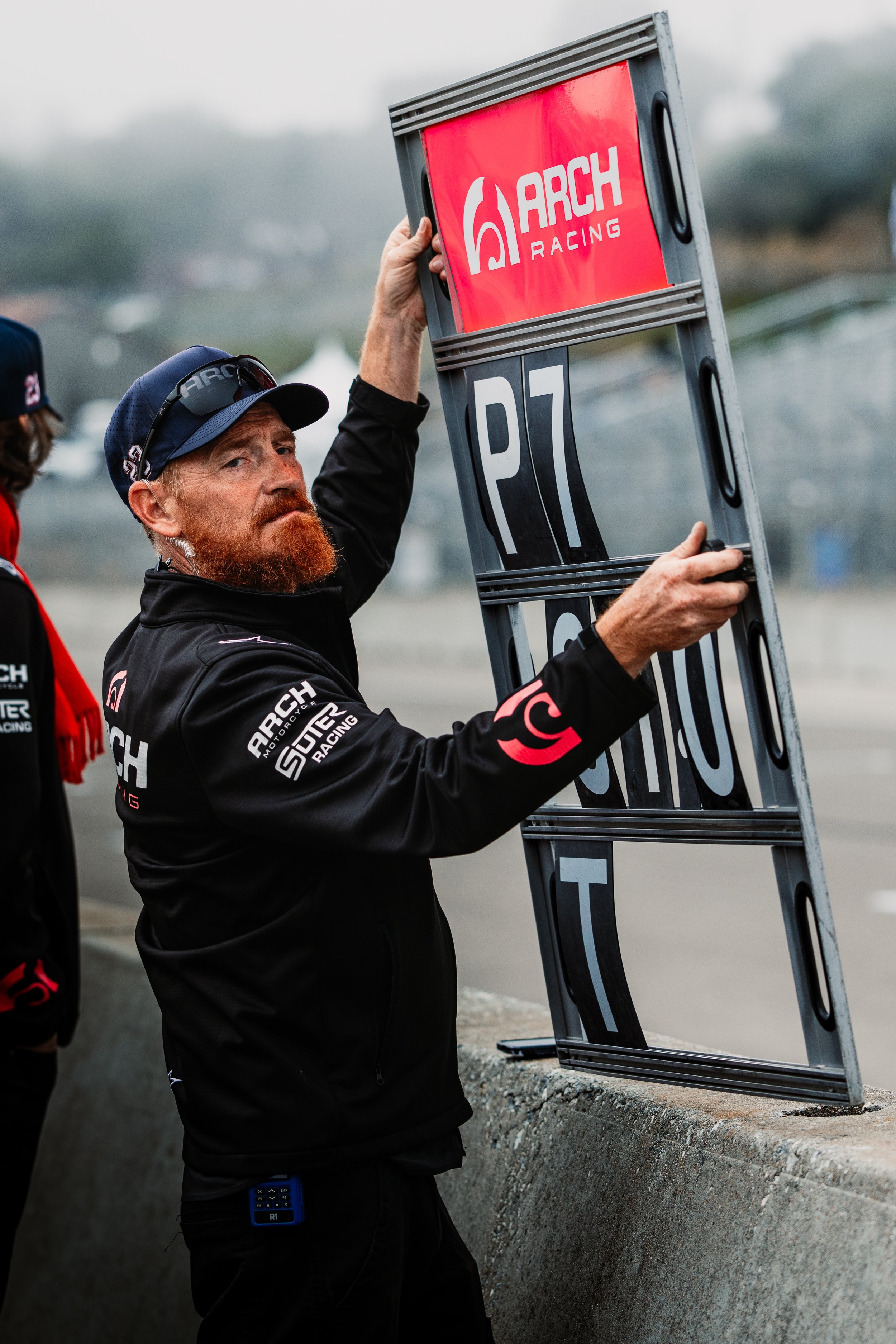 A man with a red beard and wearing a black jacket with ARCh Racing logos is holding a sign that reads 'P7' at a racing event. Another person is partially visible to the left.
