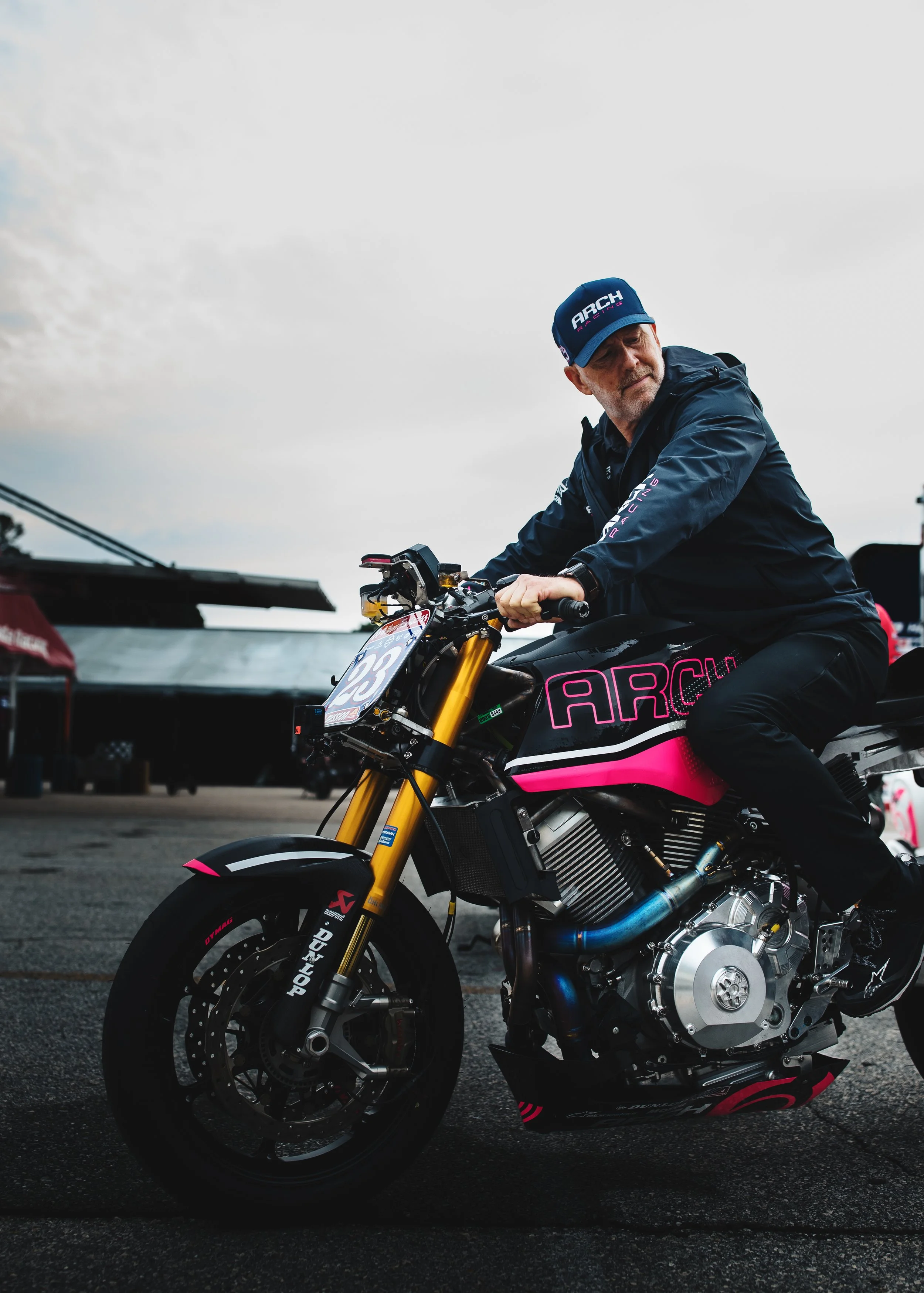 Man in black jacket and blue cap sitting on a black and pink ARC motorcycle, with yellow front forks, black tires, and visible engine, outdoors on a cloudy day.
