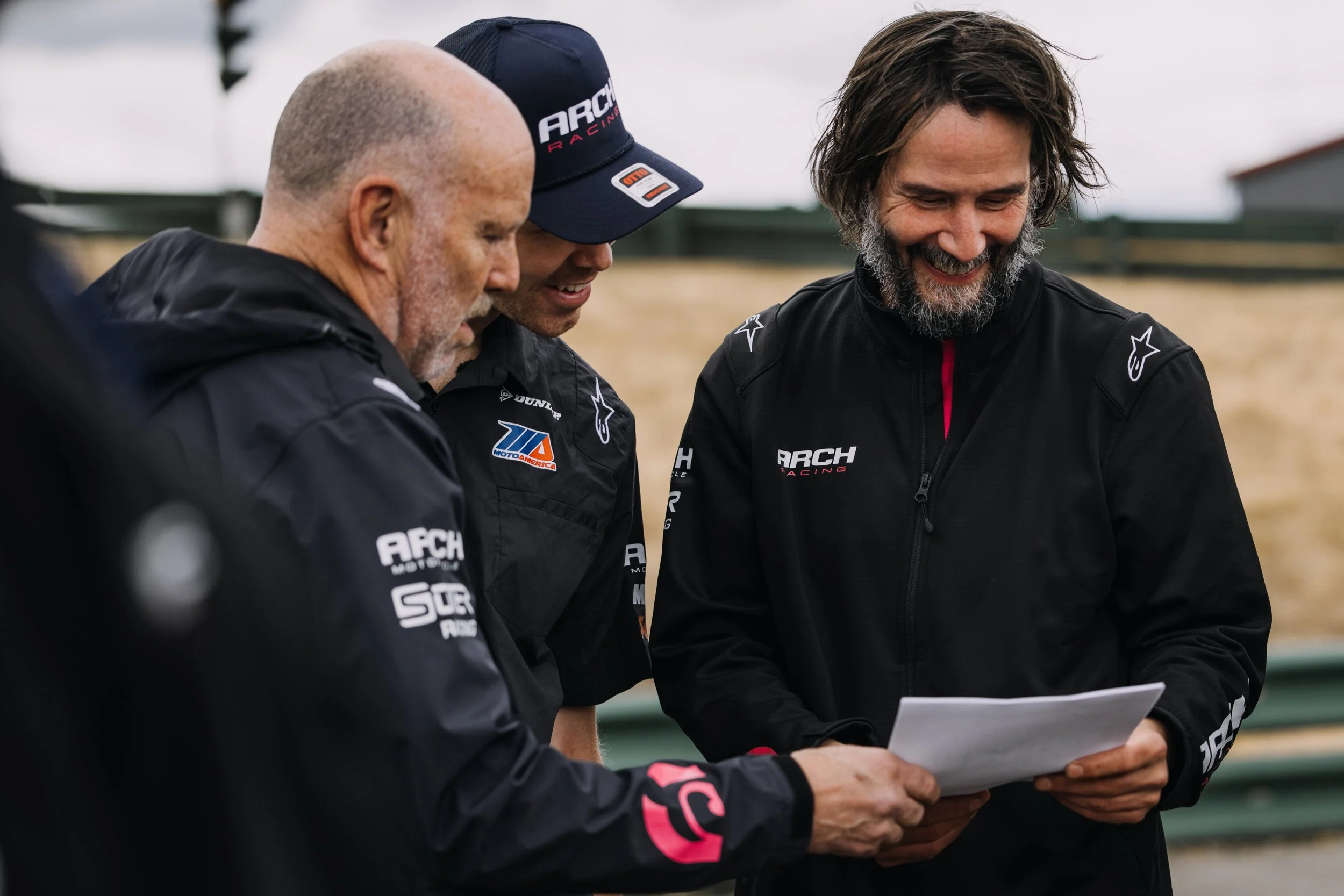 Three men in racing jackets and one wearing a cap are looking at a paper, smiling, outdoors at a racing track.