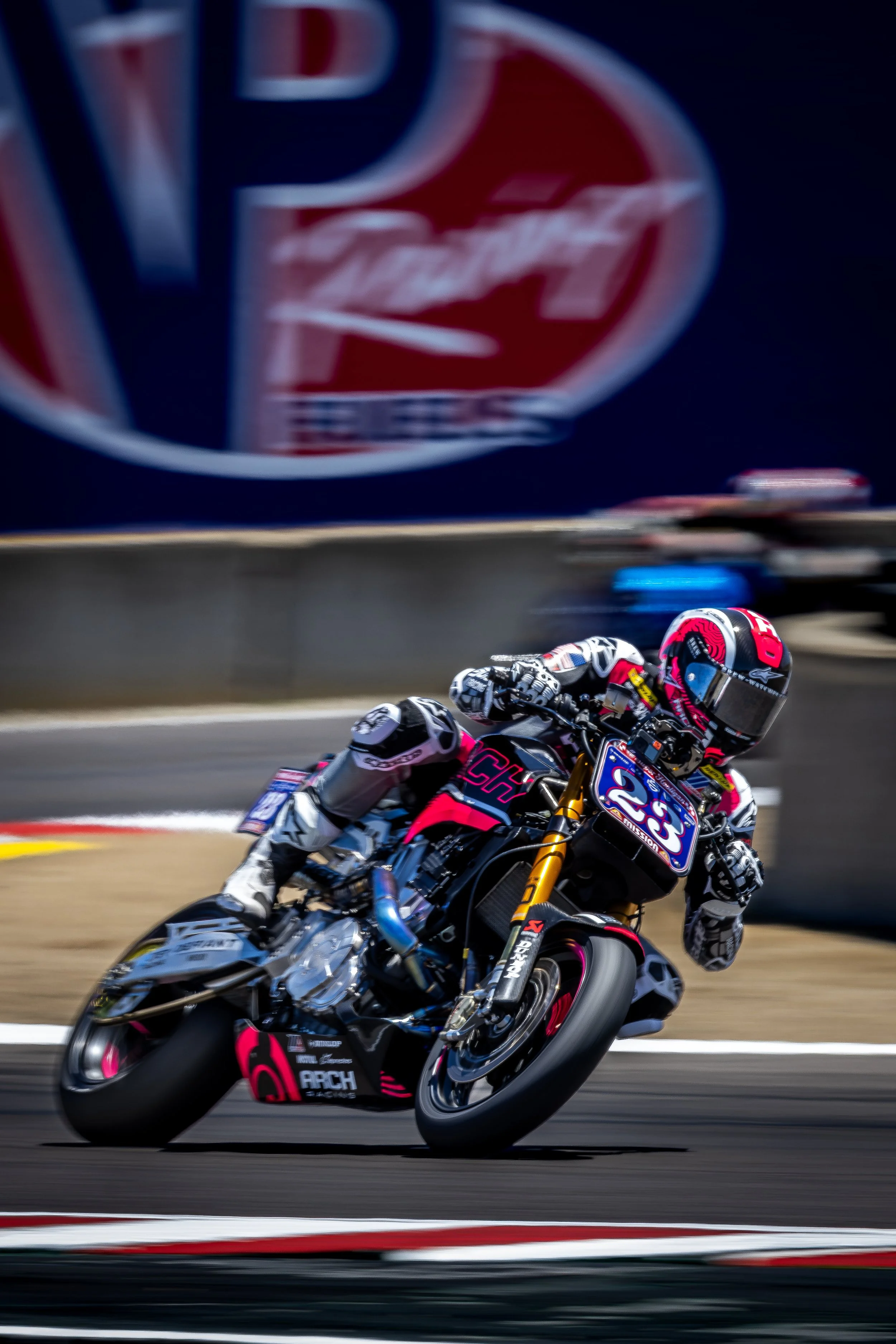 Racing motorcycle with a rider in a pink, black, and white suit leaning into a turn on a race track, with a large blurred logo of a brand in the background.