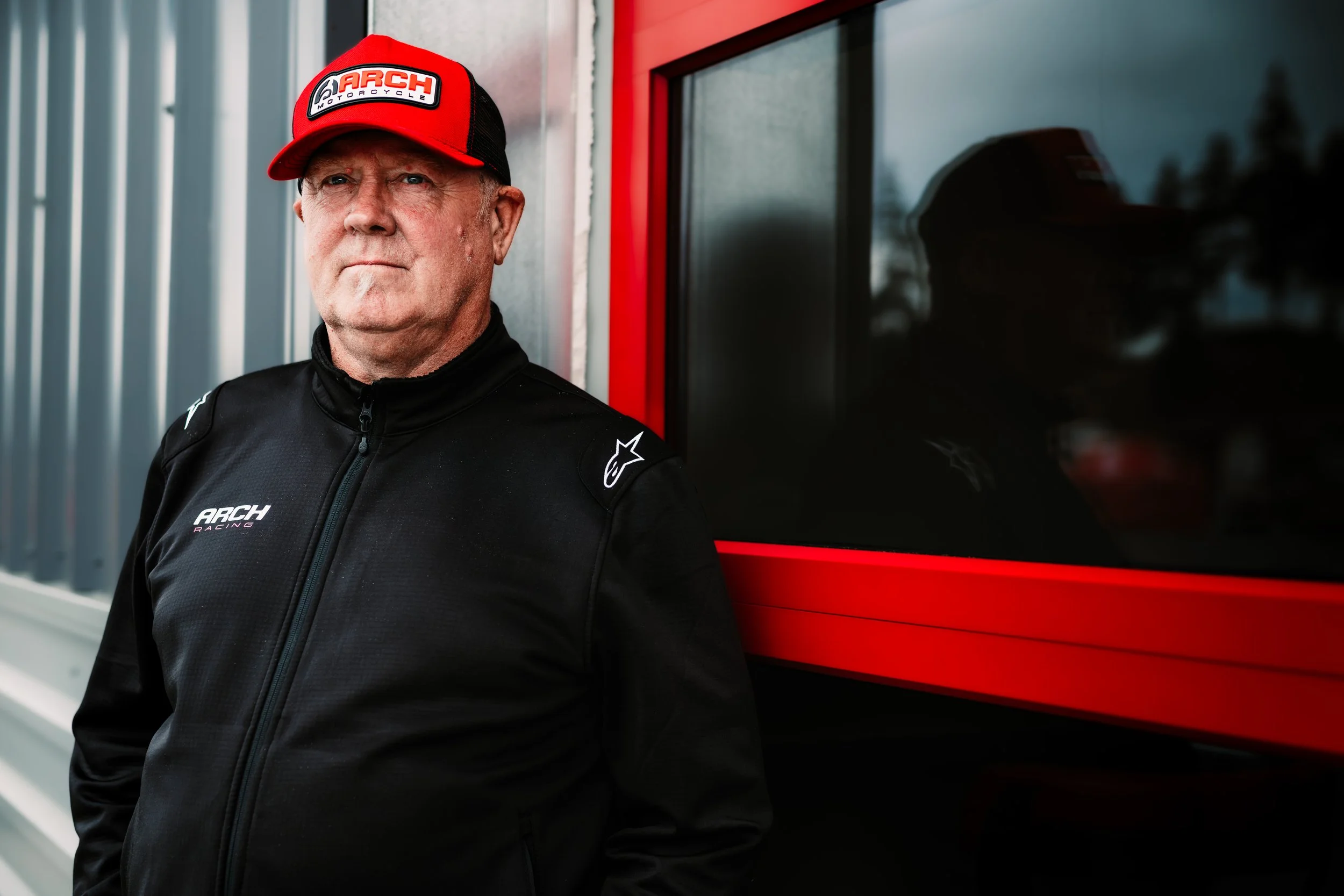 An older man in a black racing jacket and red cap standing next to a black and red structure, with a reflective window in the background.